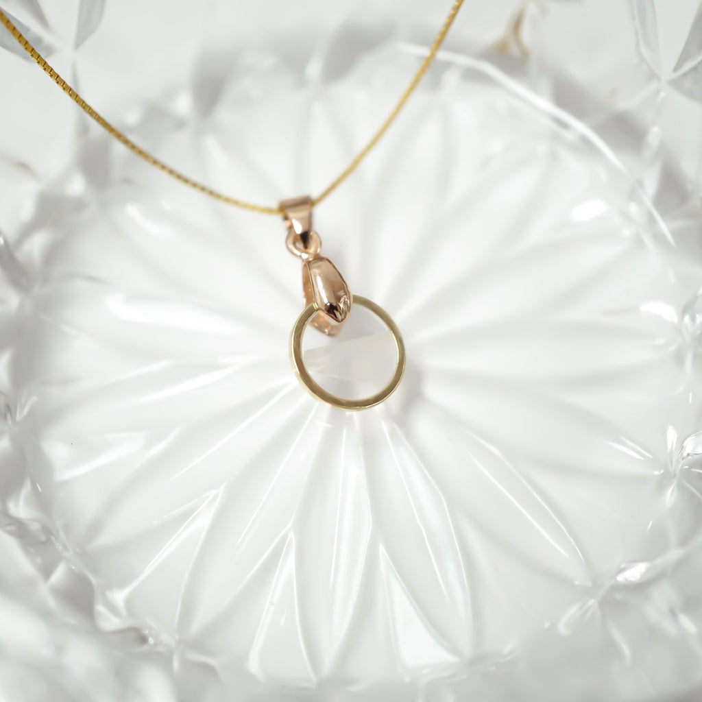 A delicate gold necklace with a small teardrop-shaped pendant and a thin circular ring, displayed on a white textured glass plate.