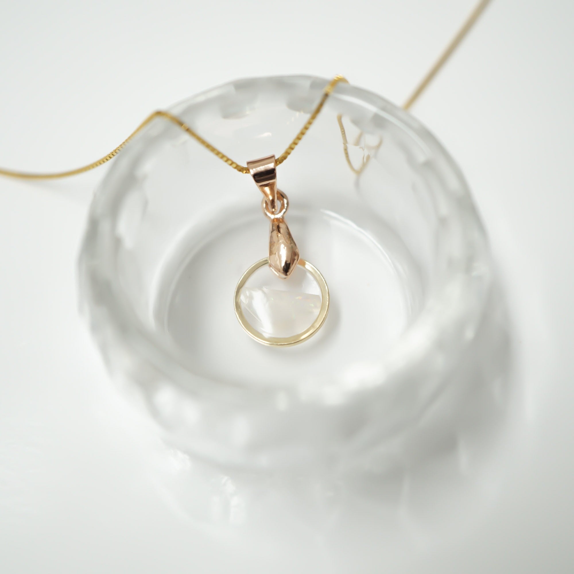 A delicate gold chain necklace with a rose gold pendant featuring a clear glass ring, displayed inside a textured clear glass dish on a white background.