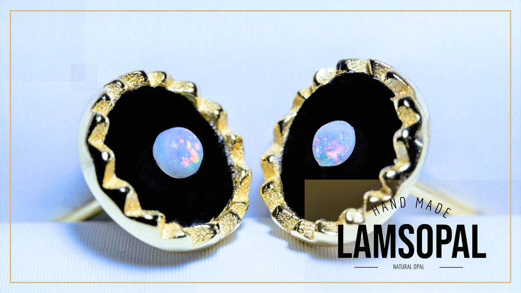 A pair of gold-toned Lamsopal cufflinks with round black inlays featuring iridescent natural opal centers, displayed on a light blue background with a gold border.