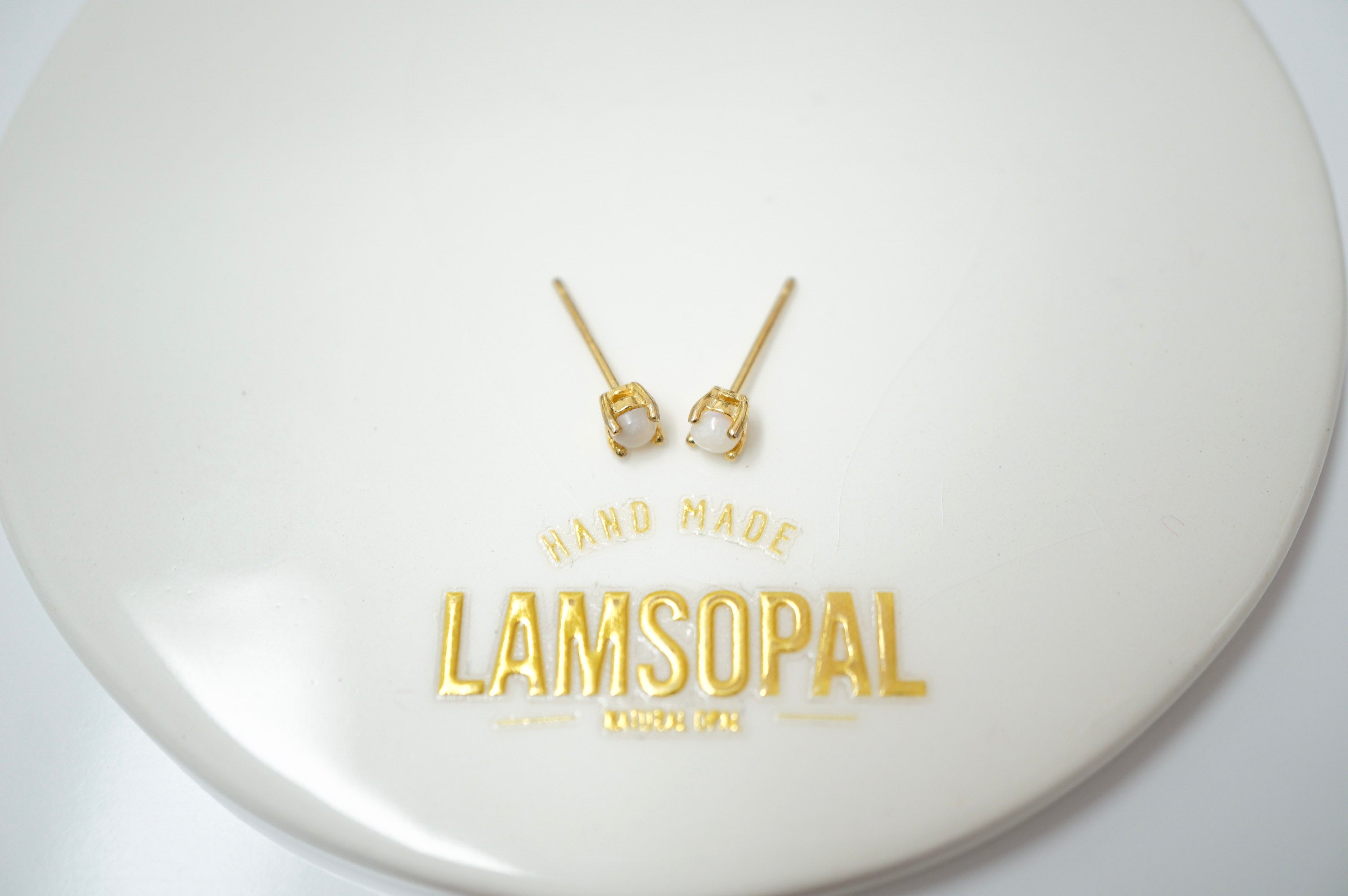 A pair of small gold stud earrings with white stone accents, displayed on a glossy white circular surface printed with the brand name 'LAMSOPAL'.
