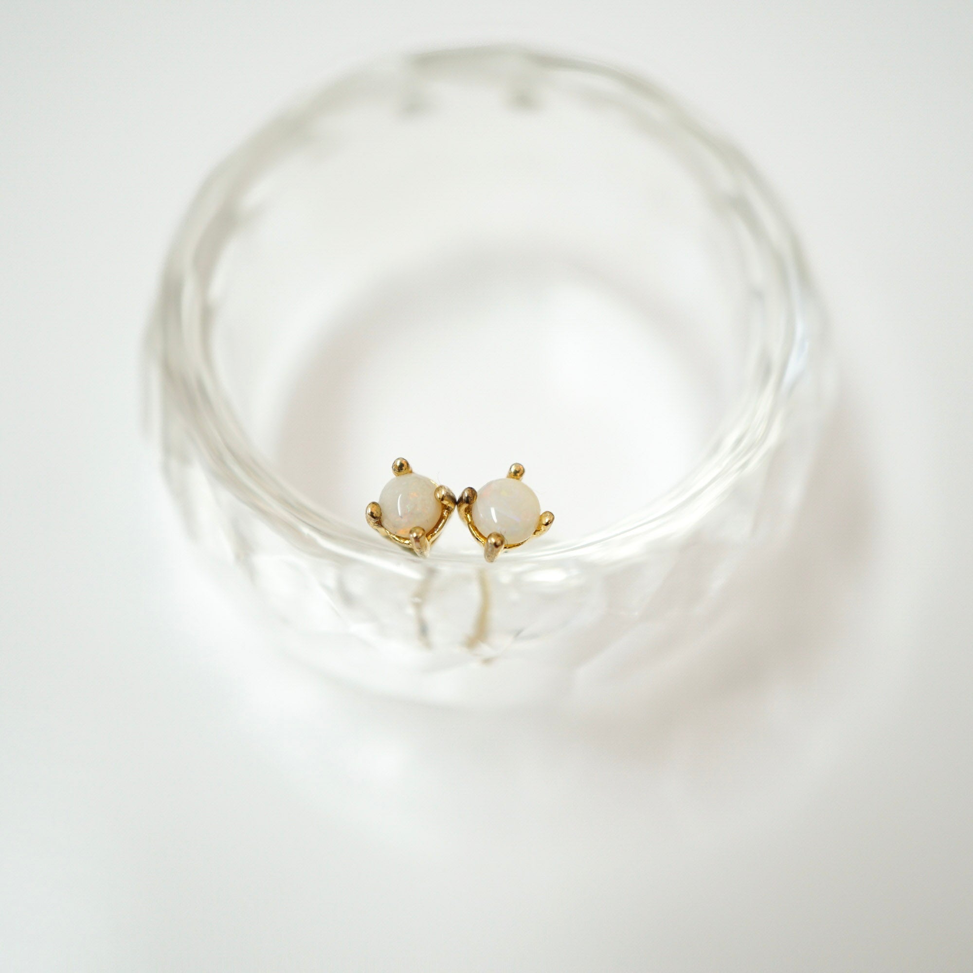A pair of small gold stud earrings with oval white stones, displayed inside a clear textured glass bowl on a white background.