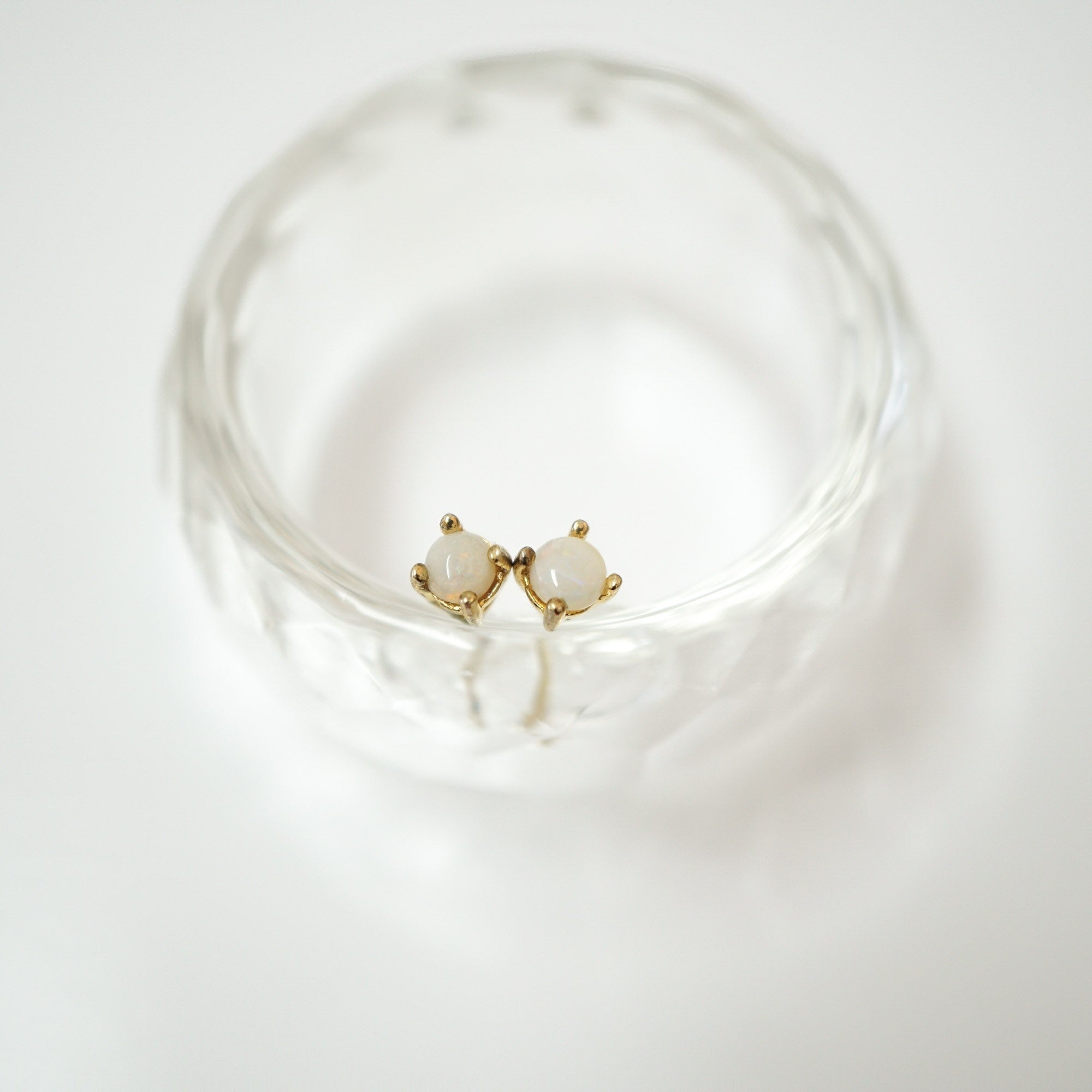 A pair of small gold stud earrings with oval white stones, displayed inside a clear textured glass bowl on a white background.