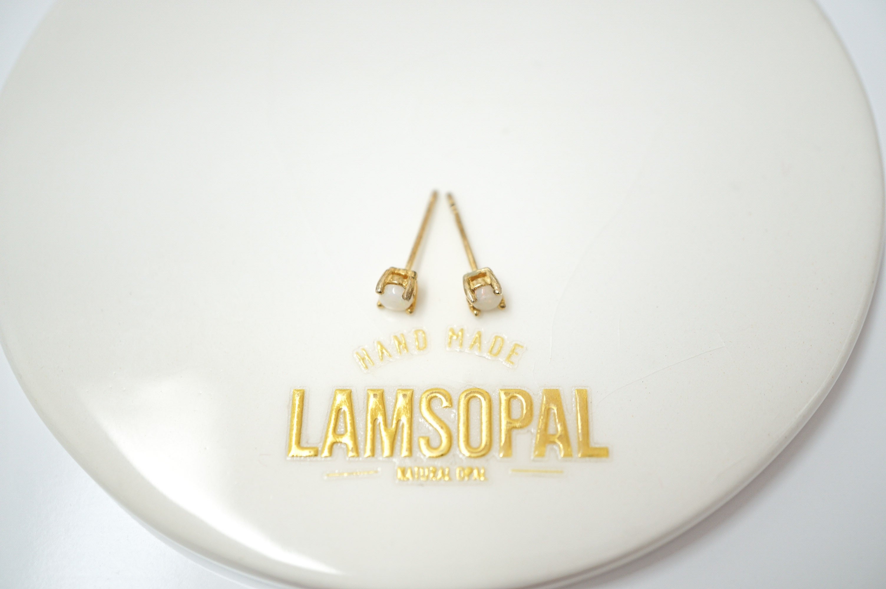A pair of gold-toned stud earrings with white pearl-like centers, displayed on a glossy white circular surface with gold foil branding that reads 'LAMSOPAL'.
