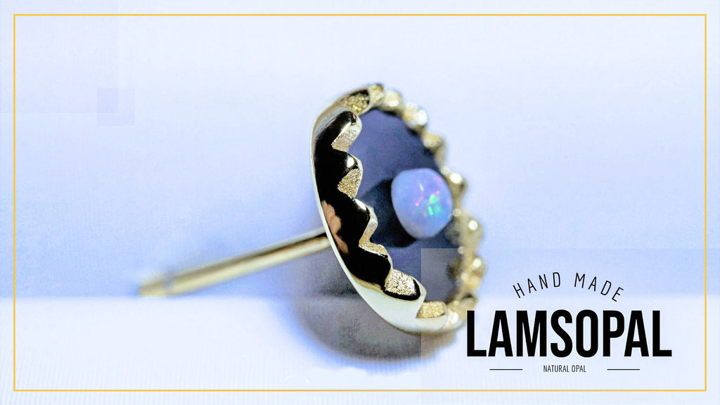 A handmade Lamsopal stud earring featuring a natural opal in a bezel setting with gold-plated detailing, shown on a light blue background with a gold border and text overlay.