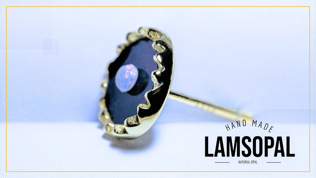 A handmade Lamsopal stud earring featuring a scalloped gold bezel holding a dark black opal with a subtle blue luster, set against a light blue background with a gold border and a brand logo overlay.