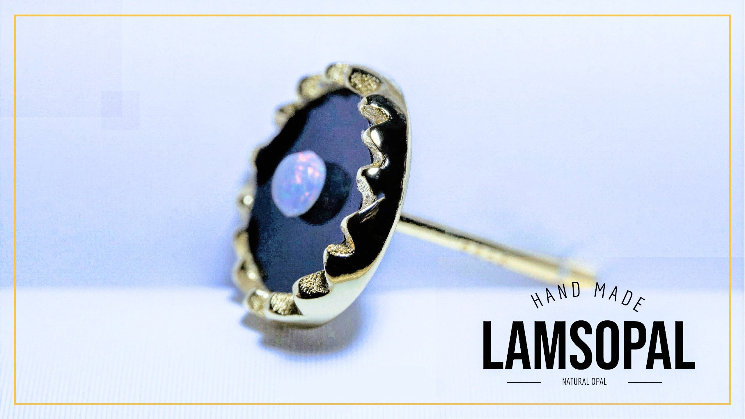 A handmade Lamsopal stud earring featuring a scalloped gold bezel holding a dark black opal with a subtle blue luster, set against a light blue background with a gold border and a brand logo overlay.