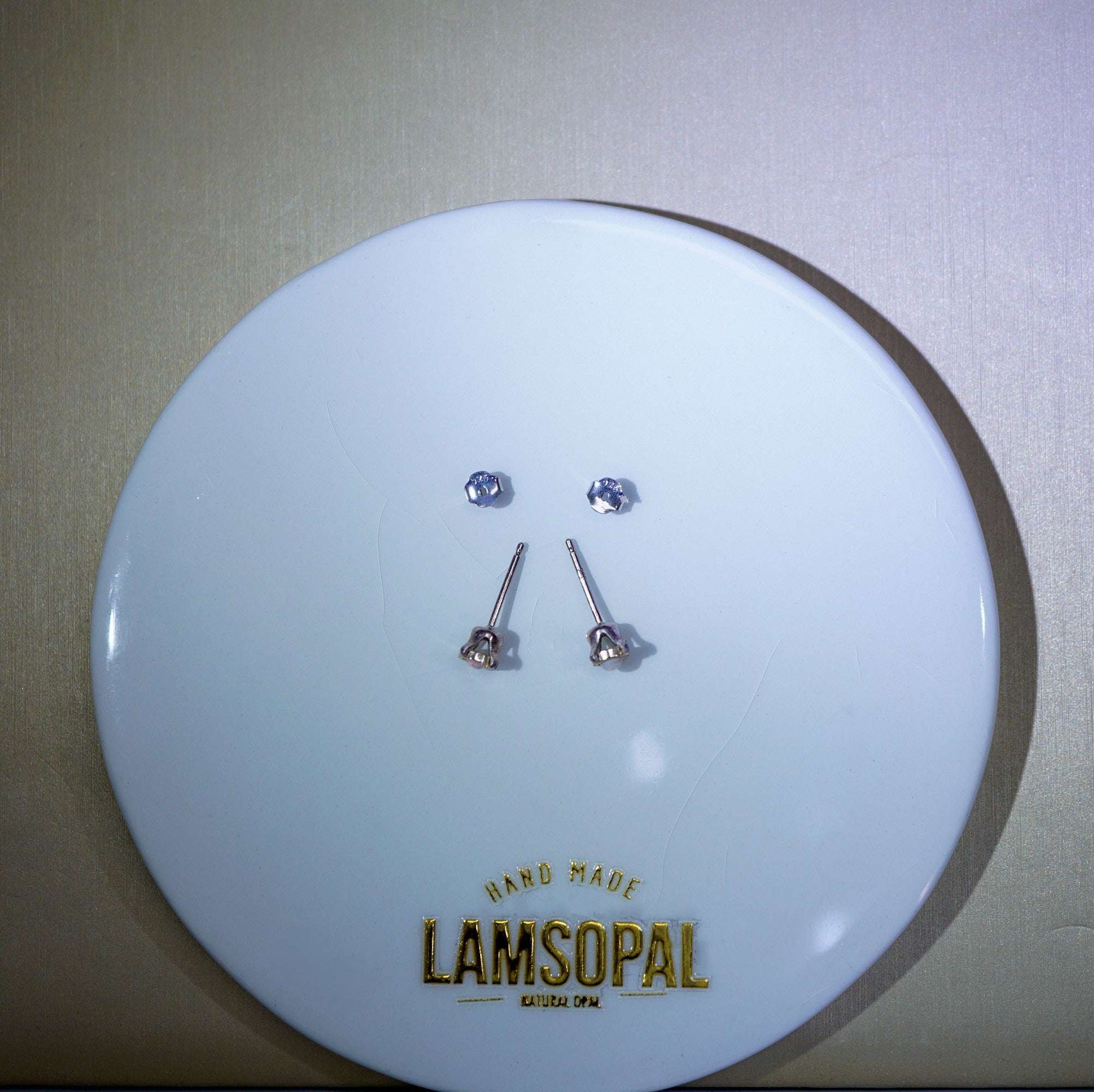 Four small stud earrings with clear faceted stones and silver posts displayed on a round white plate with the brand name LAMSOPAL printed in gold at the bottom.