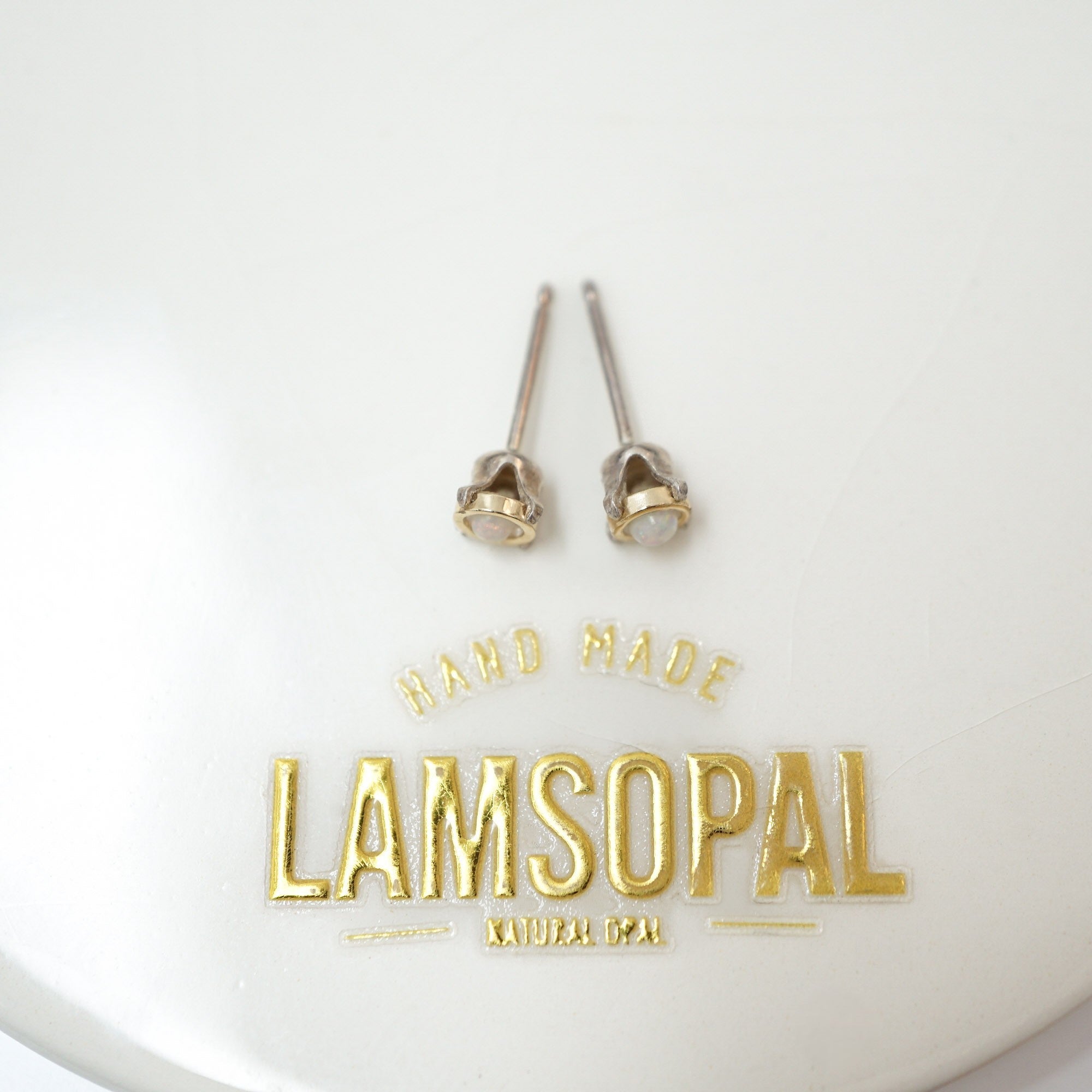 A pair of small gold stud earrings with clear gemstones in bezel settings, displayed on a white surface above a gold 'LAMSOPAL' brand logo.
