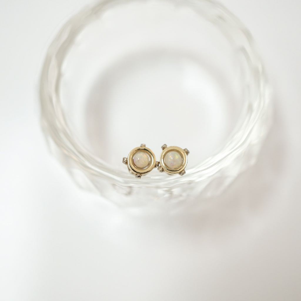 A pair of gold-toned stud earrings with oval milky white stones, resting inside a clear textured glass bowl on a white background.