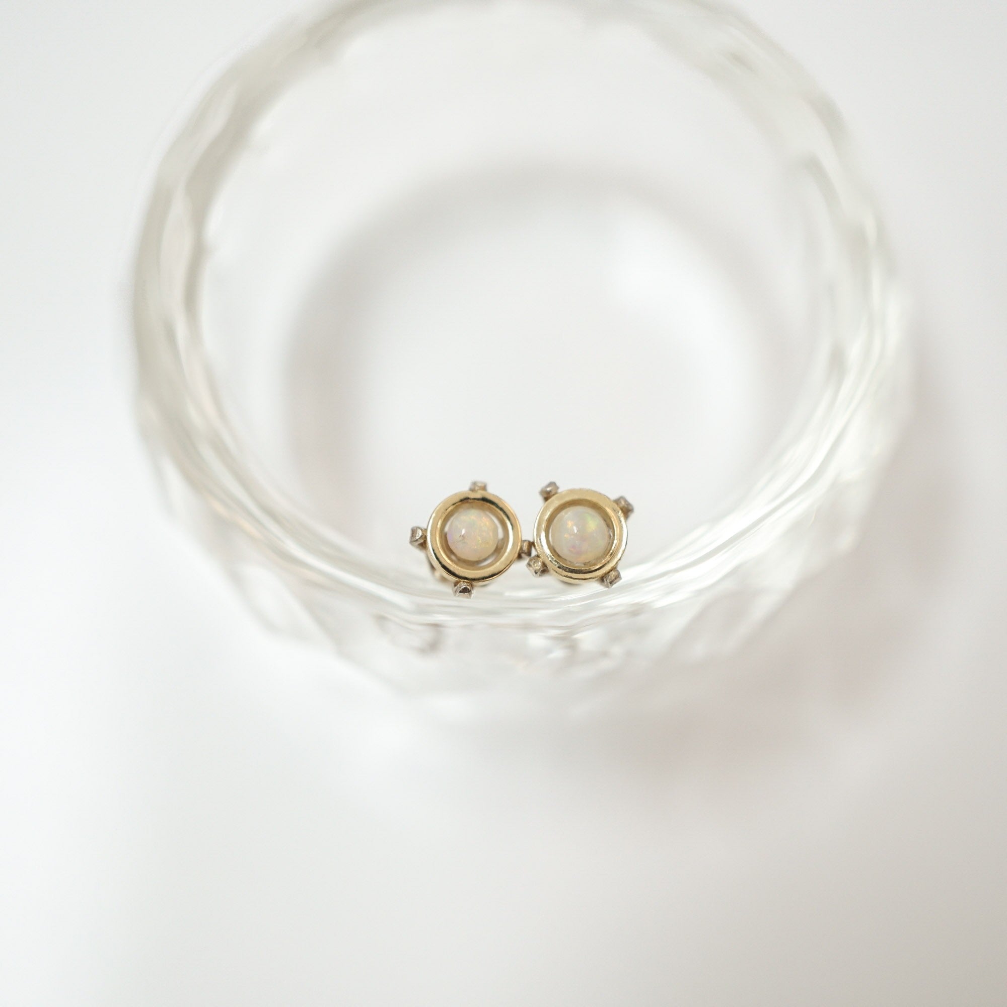 A pair of gold-toned stud earrings with oval milky white stones, resting inside a clear textured glass bowl on a white background.