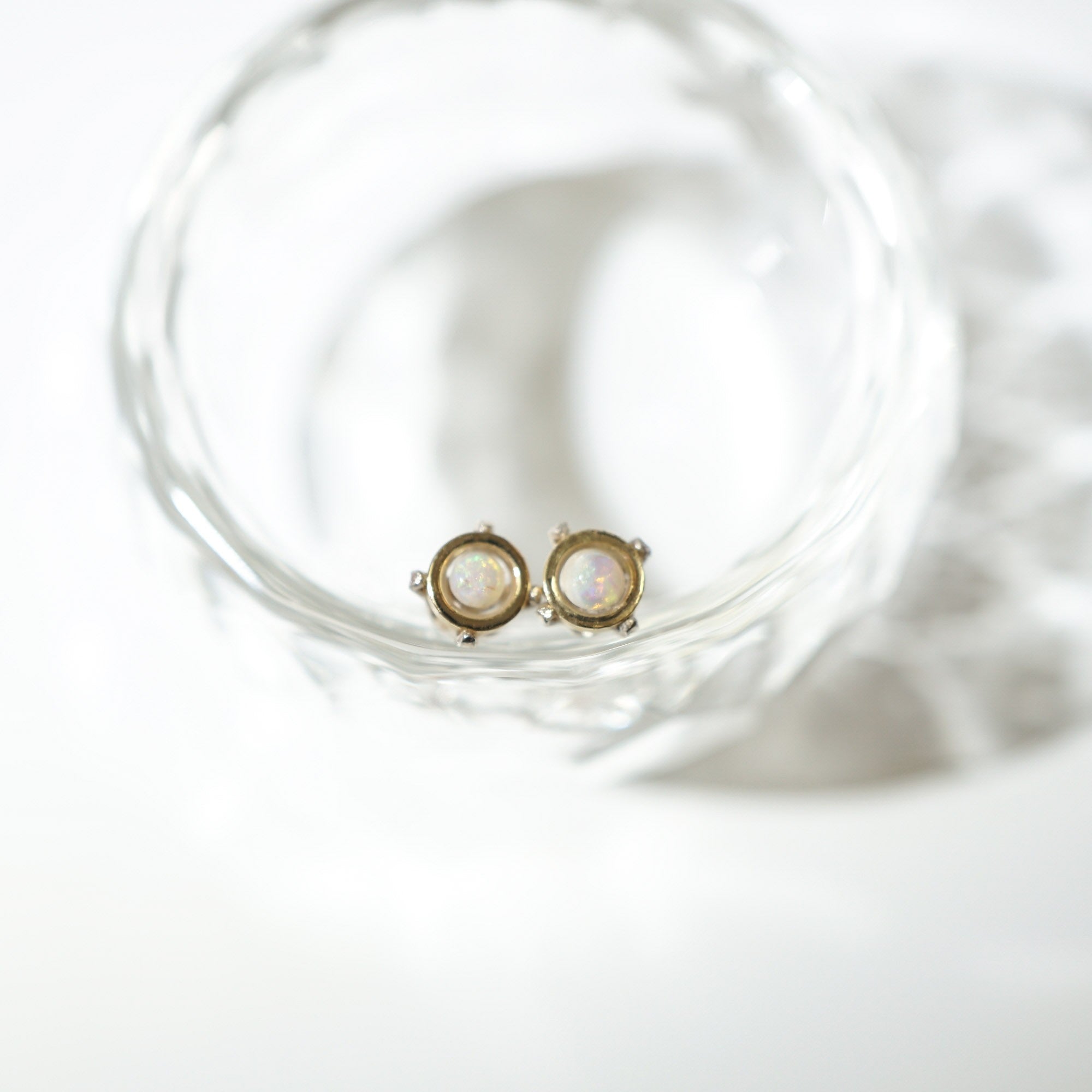 A pair of gold stud earrings with round iridescent opal-like centers, resting inside a clear textured glass bowl on a white surface.