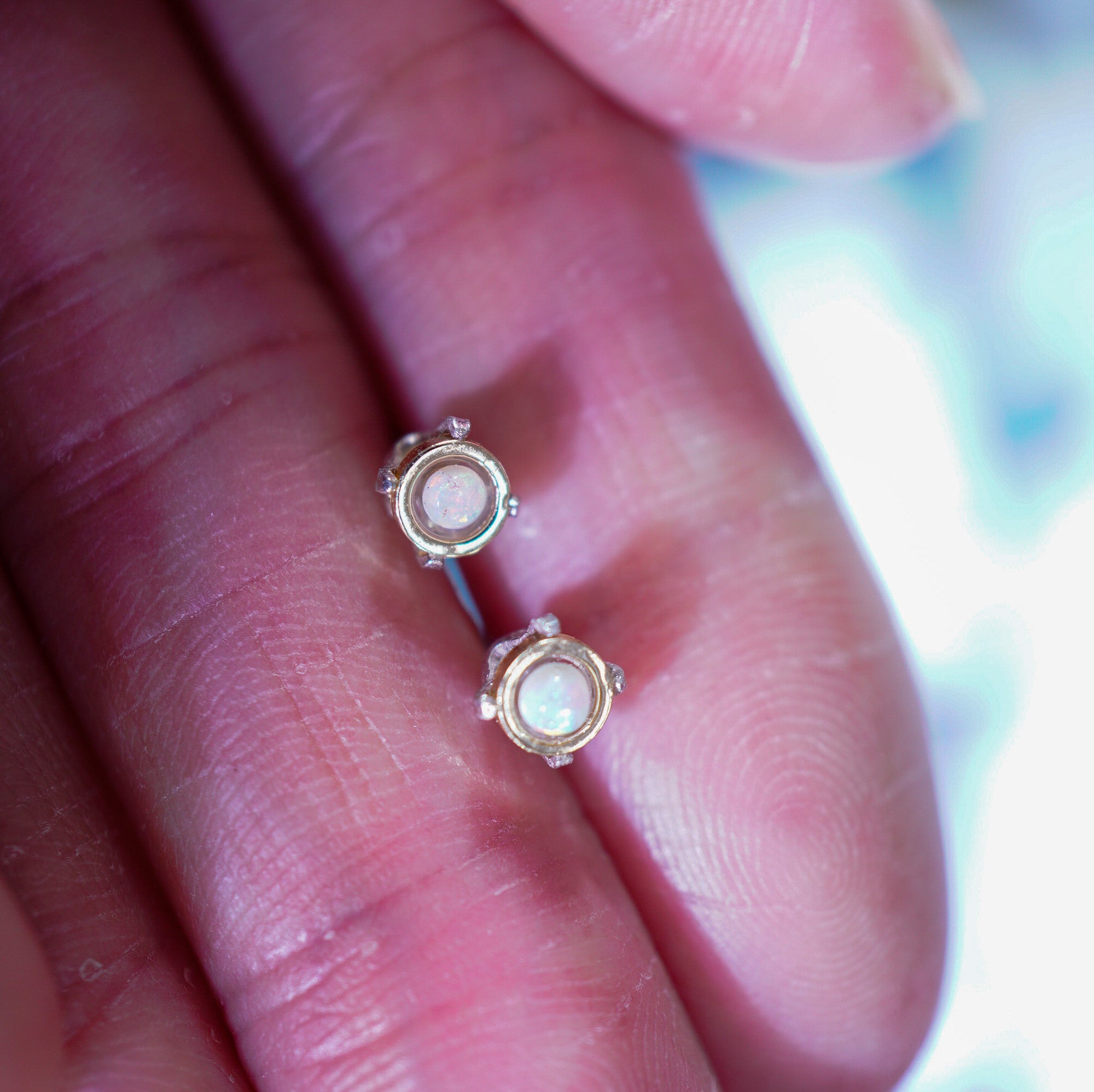 A close-up of a person's hand holding a pair of small gold stud earrings with round iridescent pinkish-white centers.