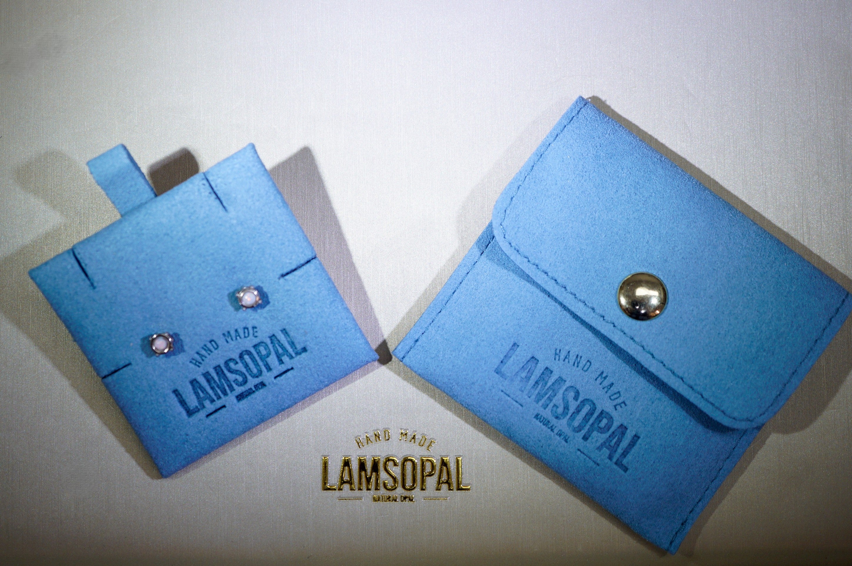 Two light blue Lamsopal card cases on a light beige background, one standing upright with a tab and two rhinestone details, and the other lying flat with a snap button closure. A Lamsopal logo watermark is visible at the bottom.