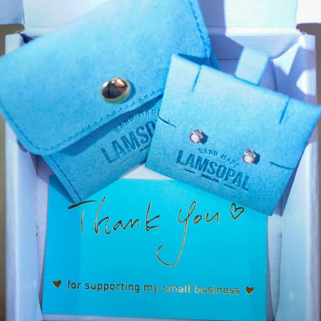 A close-up of a gift box containing two light blue velvet jewelry display cards branded LAMSOPAL, one with a snap closure and one with stud earrings, plus a turquoise thank-you card with gold script reading 'Thank you for supporting my small business'.