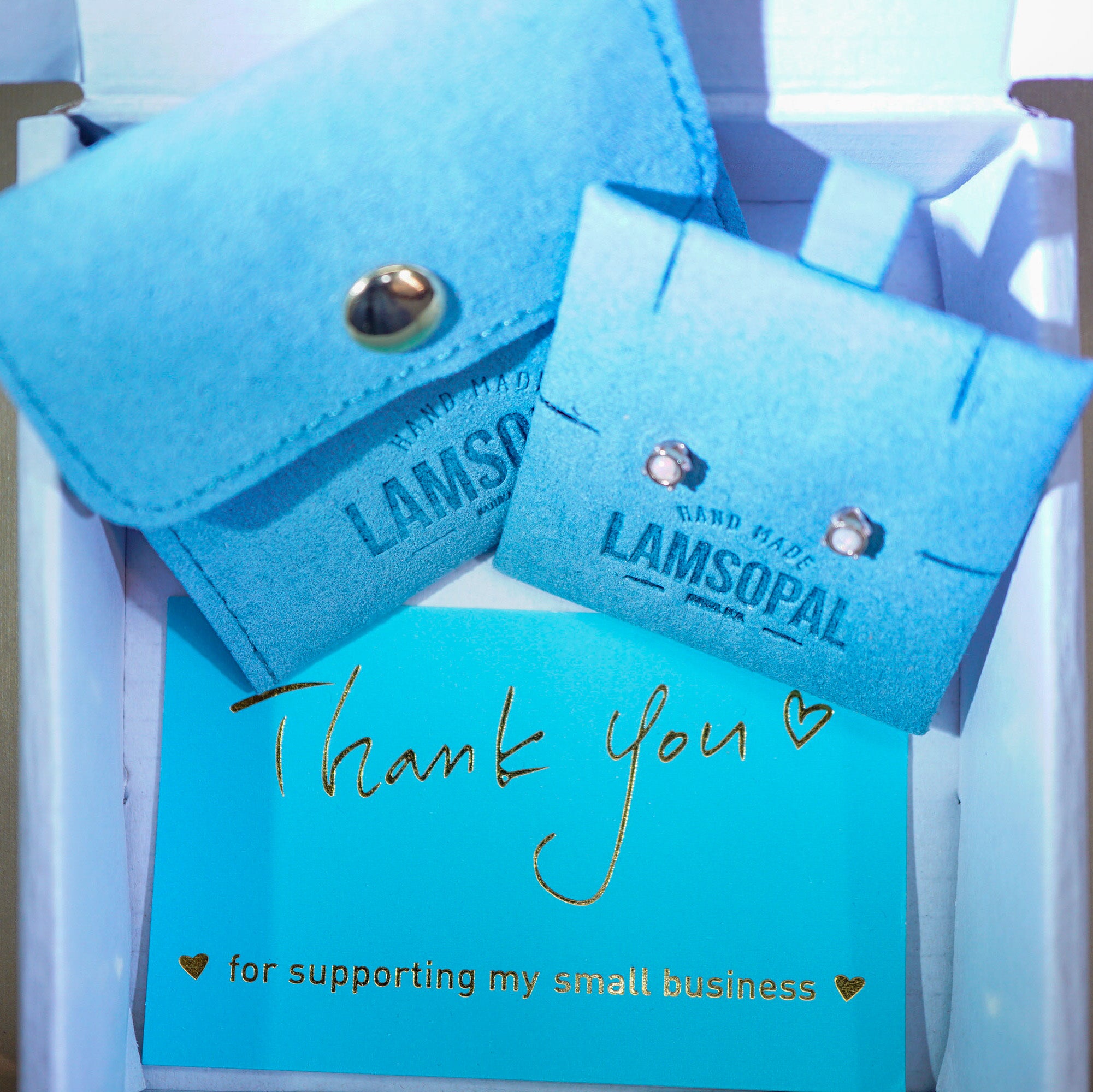 A close-up of a gift box containing two light blue velvet jewelry display cards branded LAMSOPAL, one with a snap closure and one with stud earrings, plus a turquoise thank-you card with gold script reading 'Thank you for supporting my small business'.