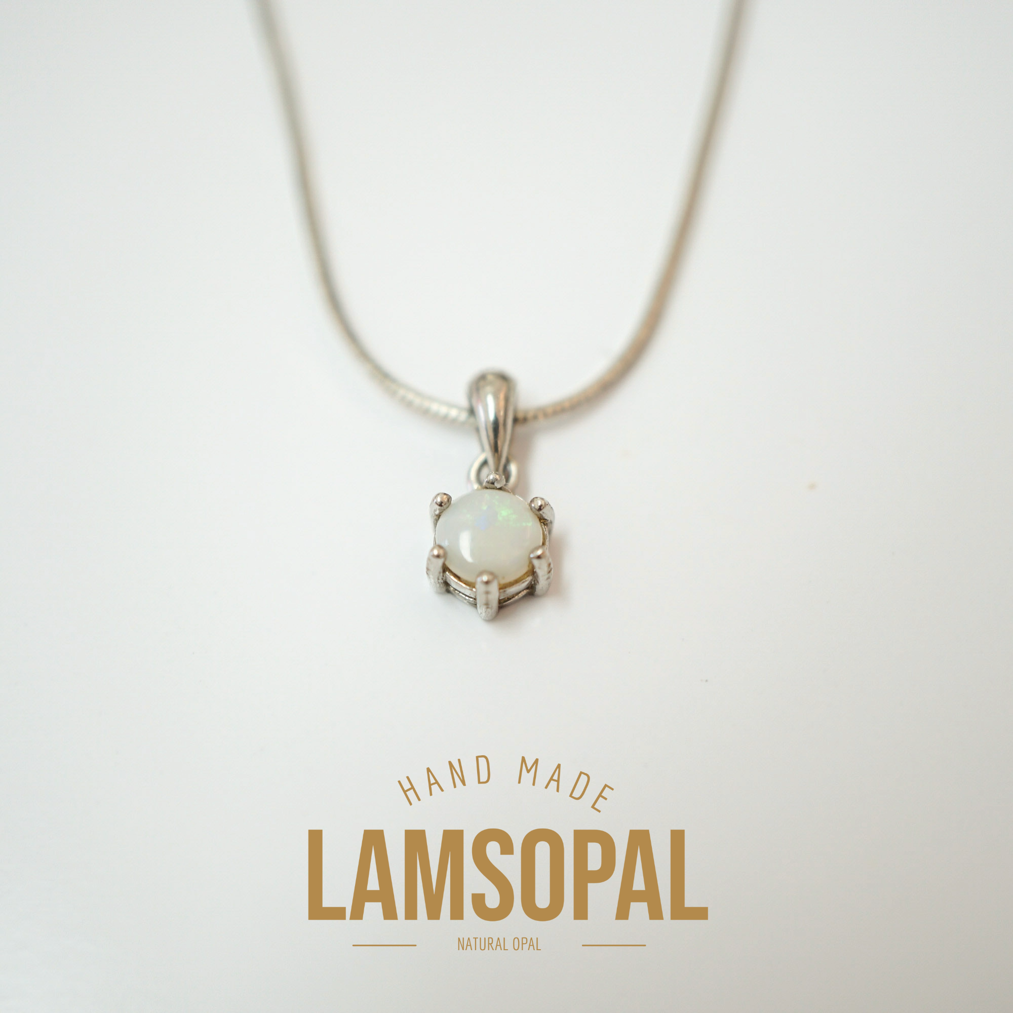 A silver necklace with a round iridescent white opal pendant in a prong setting, suspended from a silver snake chain on a white background. Below the necklace, the text 'HAND MADE LAMSOPAL NATURAL OPAL' is printed in gold.