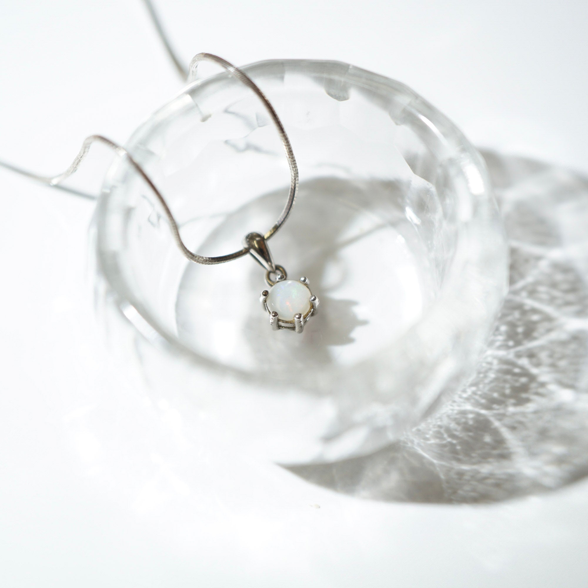 A silver-toned necklace with a small white opal-like pendant, draped over a clear glass bowl on a bright white background.