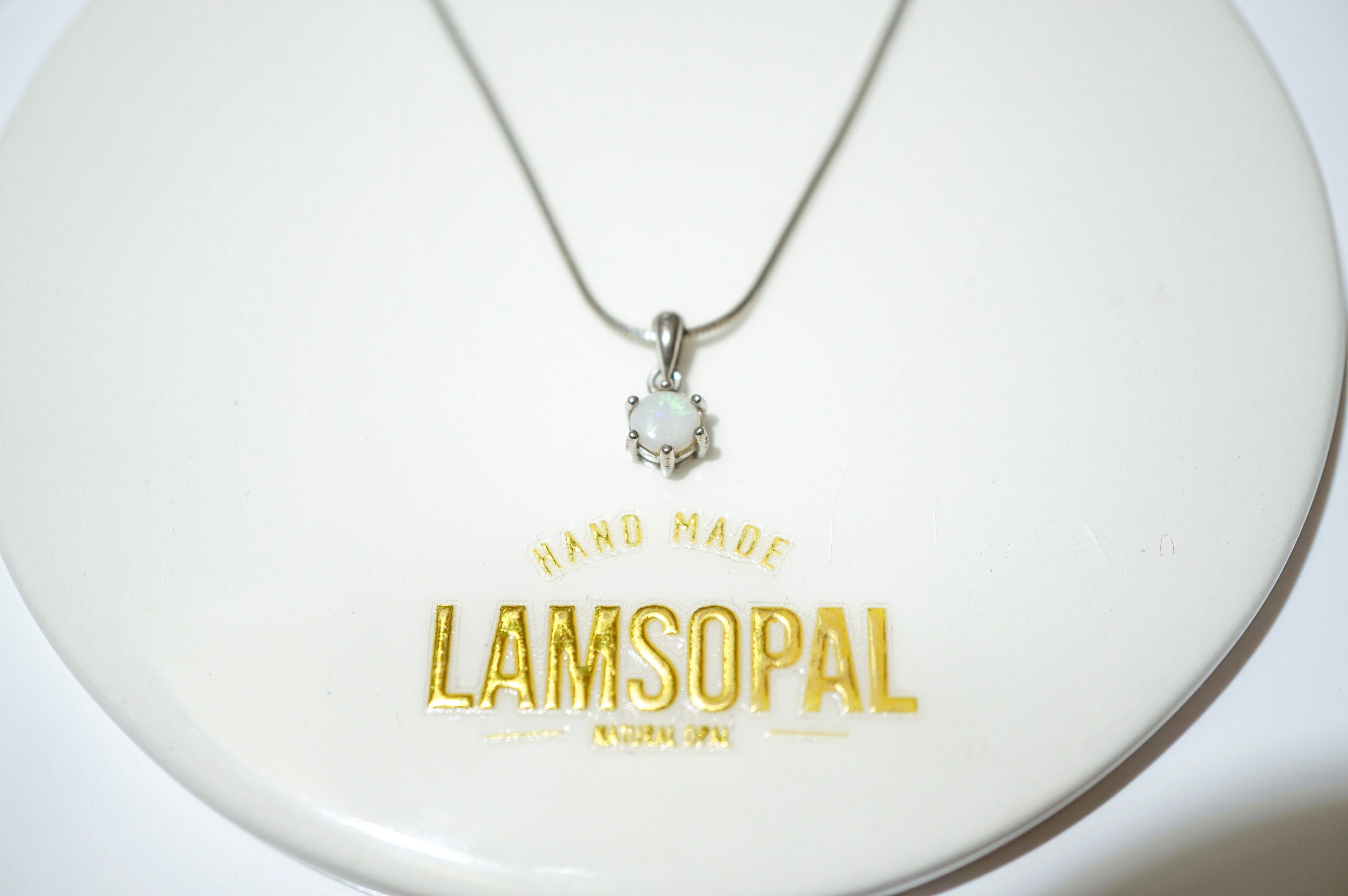 A silver necklace with a small pendant featuring a white stone, displayed on a white circular stand with gold 'LAMSOPAL' branding.
