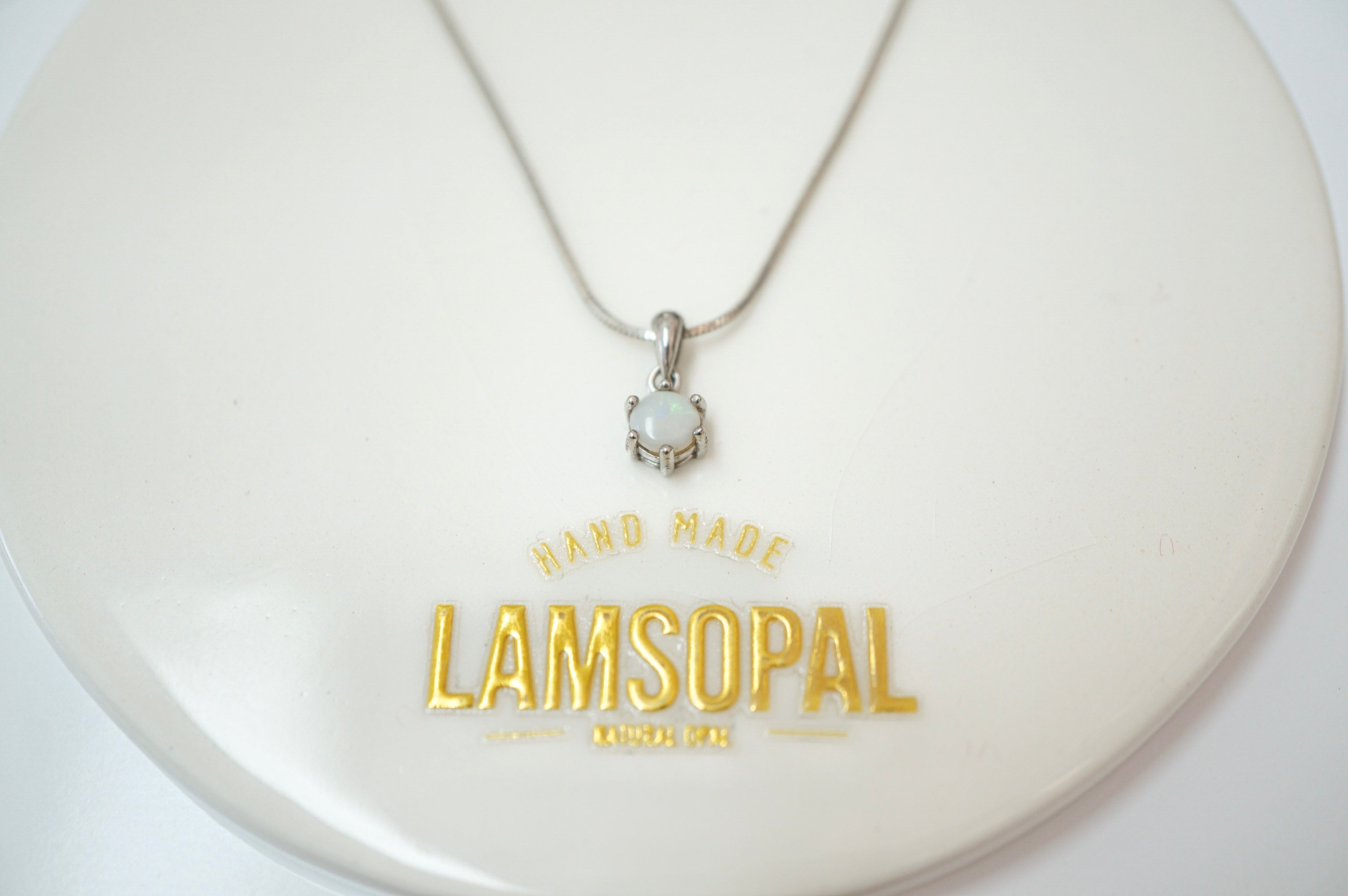 A silver chain necklace with a small silver pendant featuring a white stone, displayed on a round white stand with the brand name 'LAMSOPAL' printed in gold.