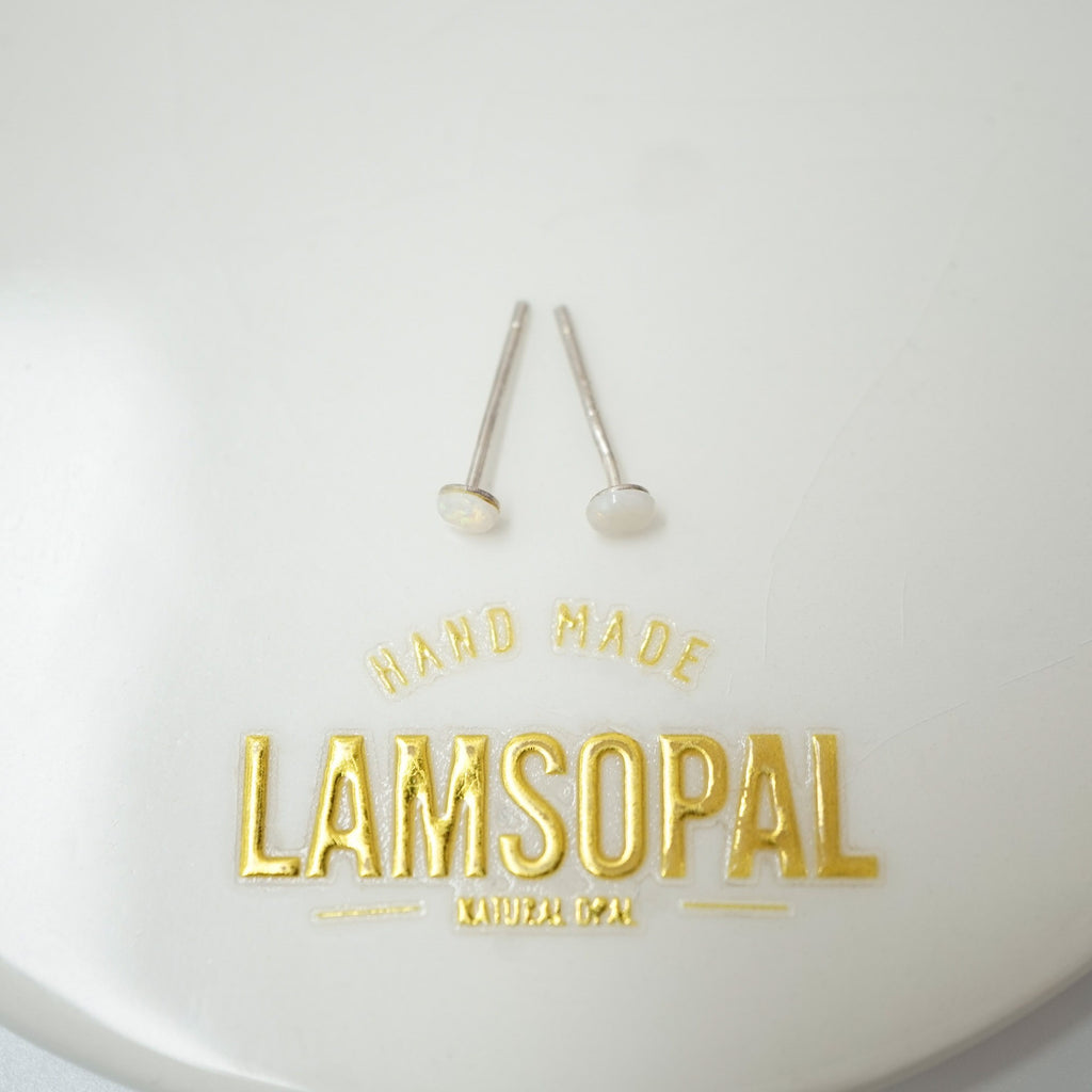 Two small silver stud earrings with round white inlay tops displayed on a white surface above gold foil branding that reads 'HAND MADE LAMSOPAL NATURAL GOOP'.