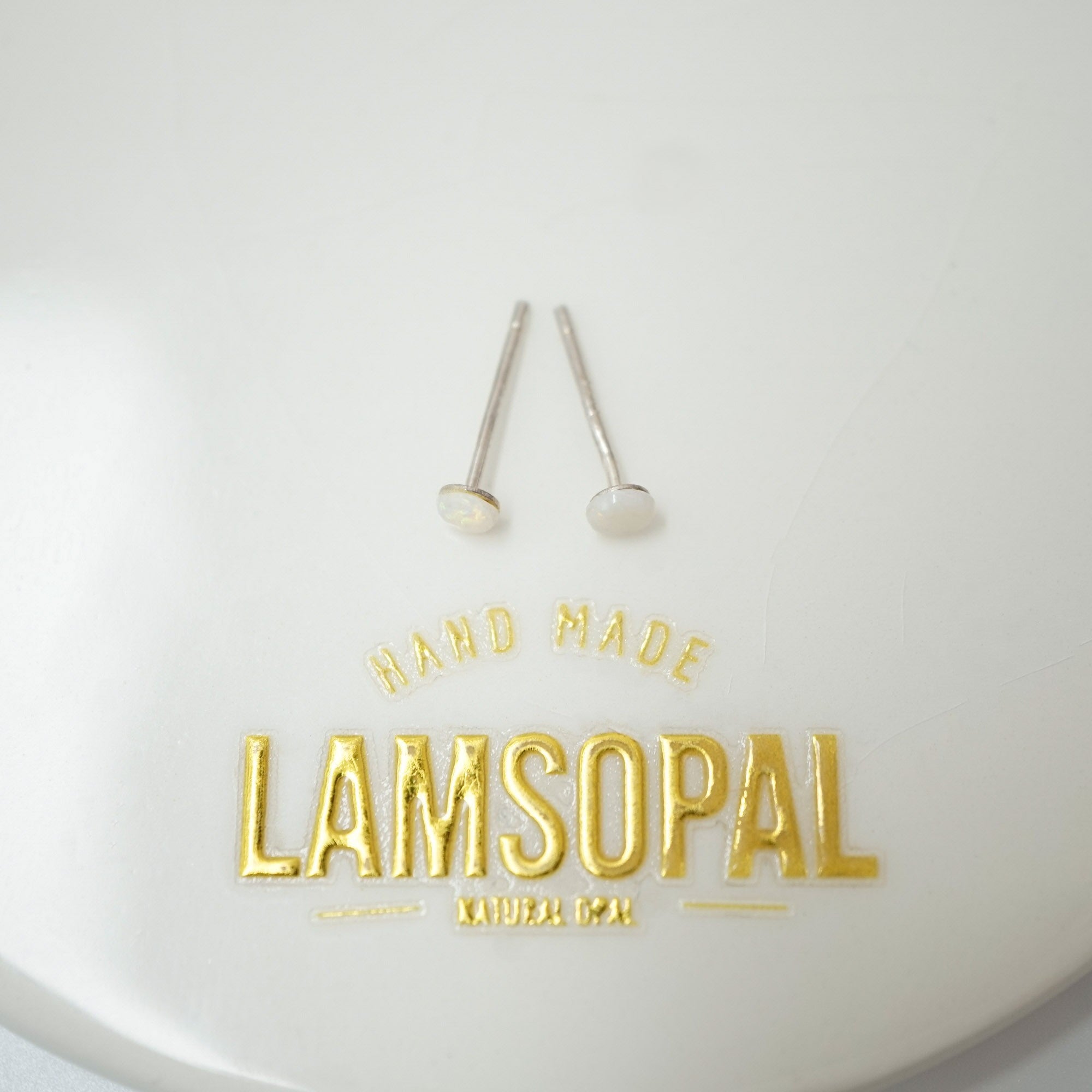Two small silver stud earrings with round white inlay tops displayed on a white surface above gold foil branding that reads 'HAND MADE LAMSOPAL NATURAL GOOP'.