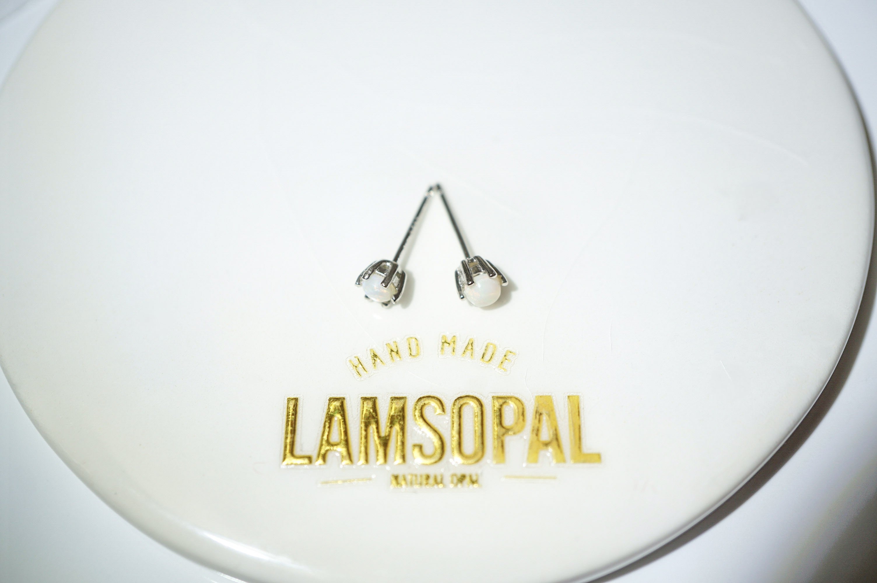 A pair of silver stud earrings with white pearl-like centers, displayed on a white circular plate printed with the brand name 'LAMSOPAL'.