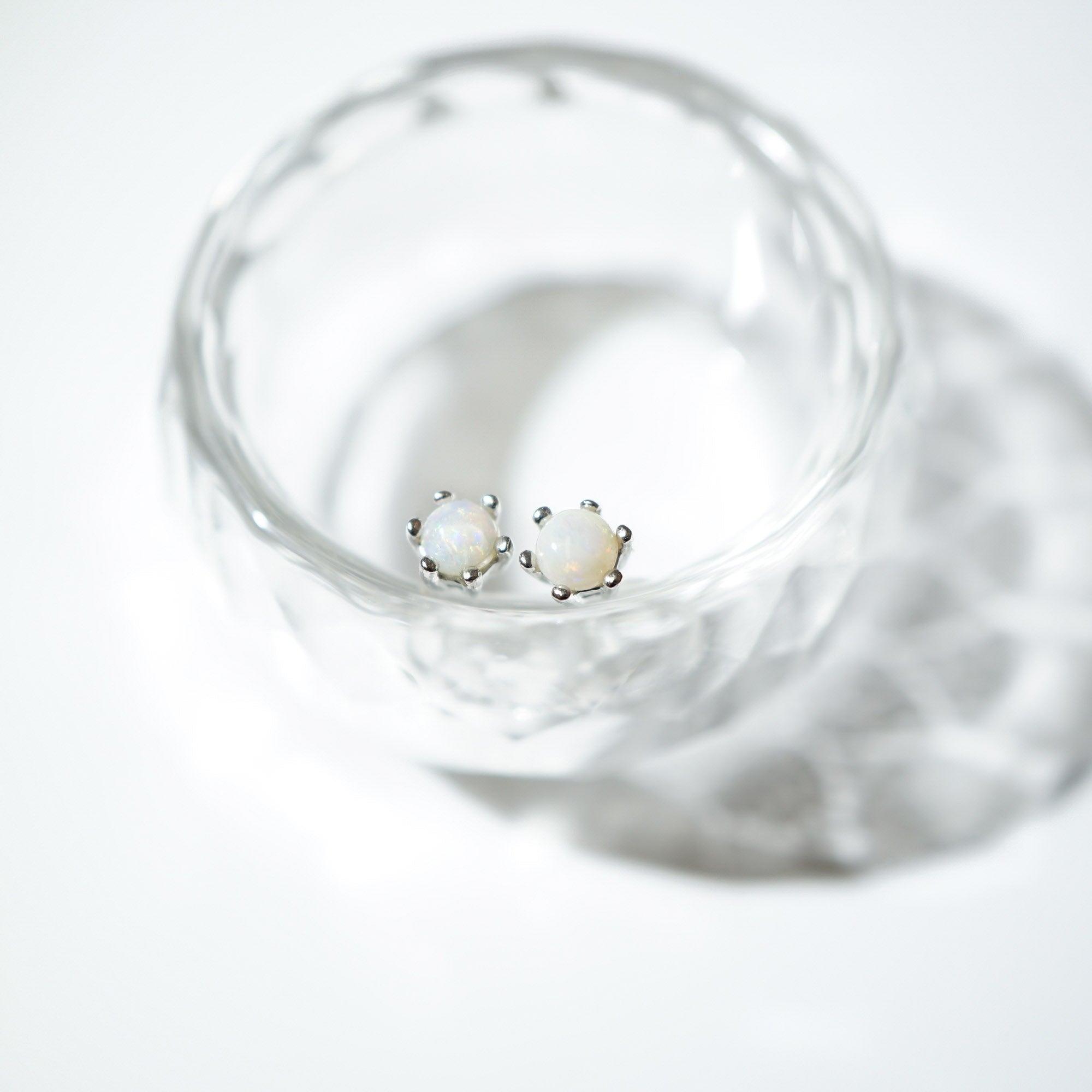 A pair of silver stud earrings with round white opal-like centers, resting inside a clear glass dish on a white background.