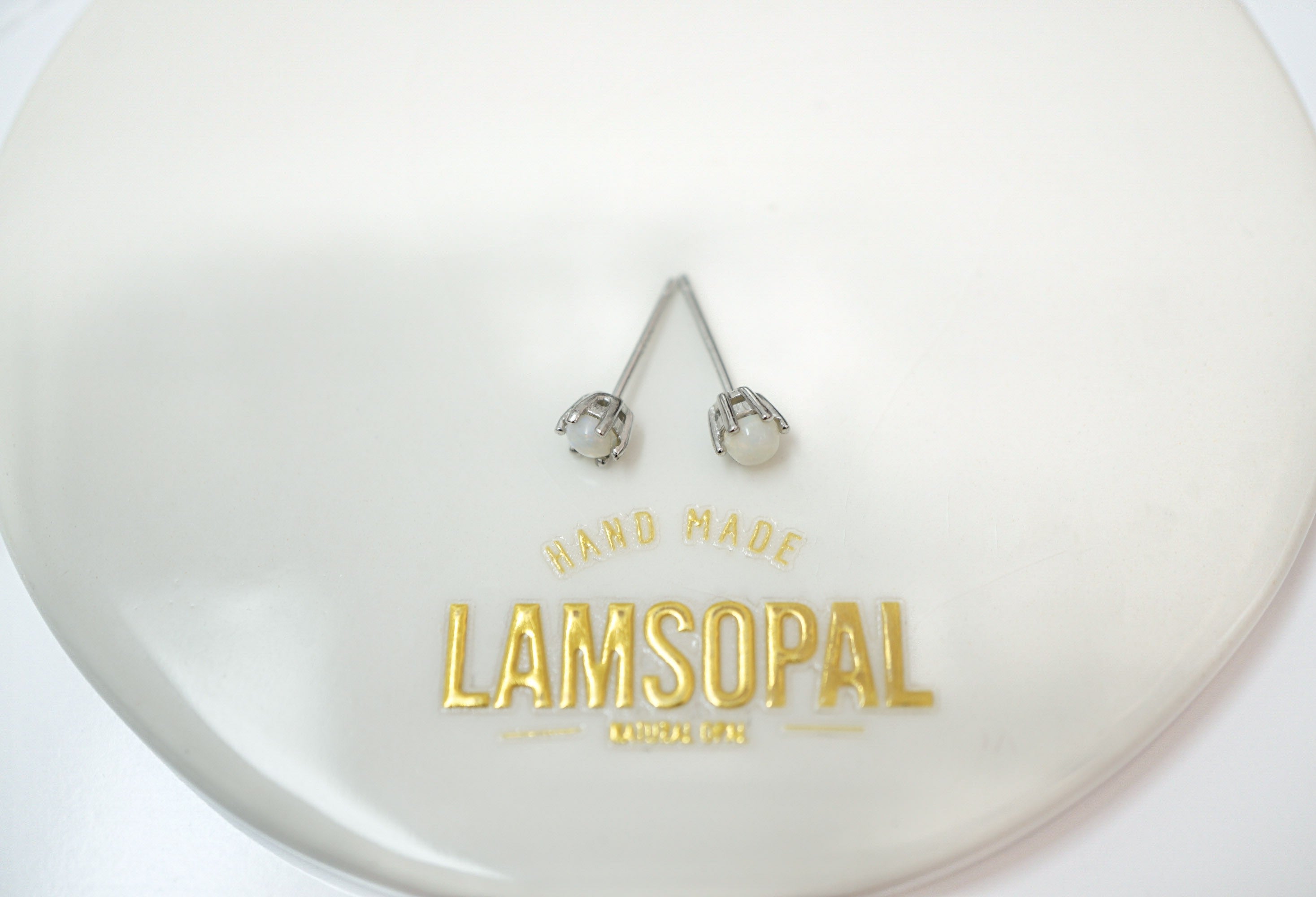 A pair of silver-toned spiral stud earrings displayed on a glossy white oval tray with gold foil branding that reads 'HAND MADE LAMSOPAL'.