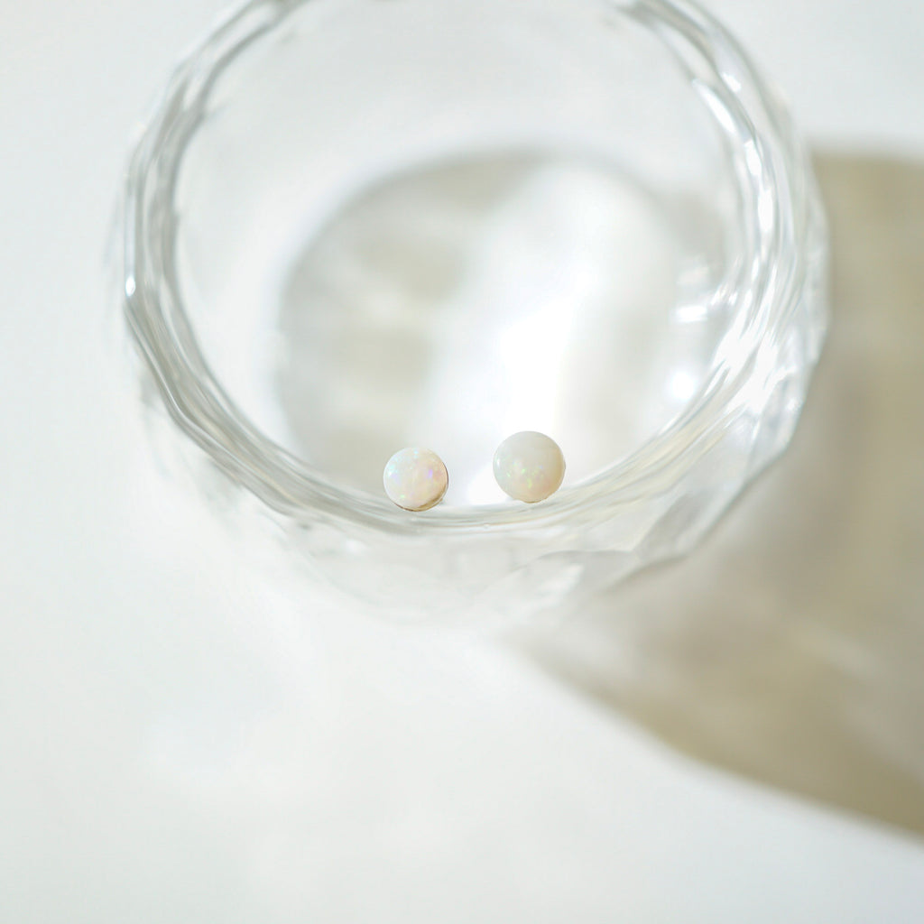 Two small round pearl stud earrings resting inside a clear textured glass bowl on a bright white surface.