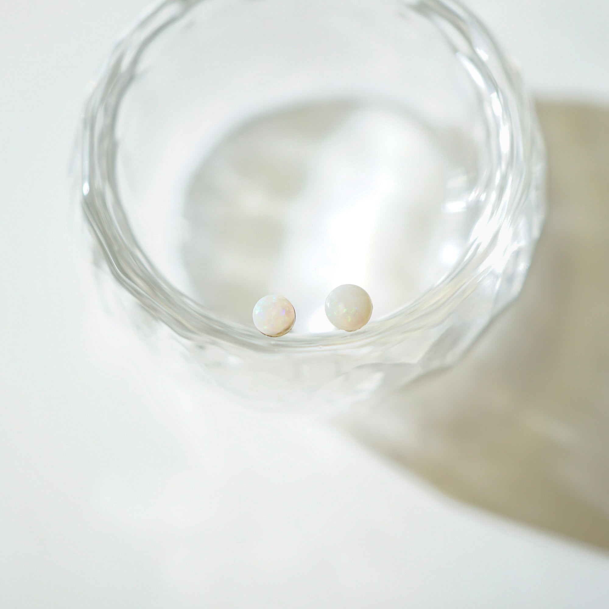 Two small round pearl stud earrings resting inside a clear textured glass bowl on a bright white surface.