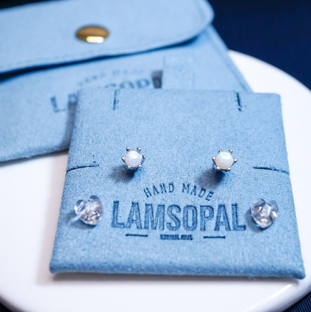 A pair of silver stud earrings with white pearl-like centers, displayed on a blue suede card branded LAMSOPAL, with additional loose clear stones and a matching blue pouch in the background.
