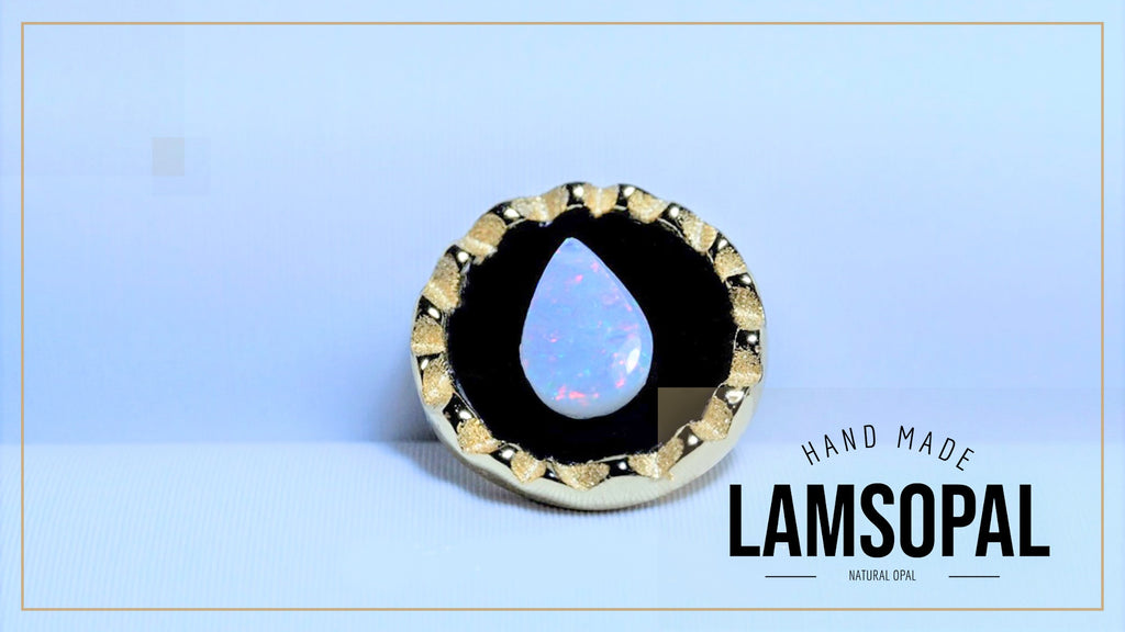 A handmade Lamsopal ring featuring a large natural opal teardrop inlay surrounded by a black and gold textured border, set against a light blue background with a brand logo overlay.