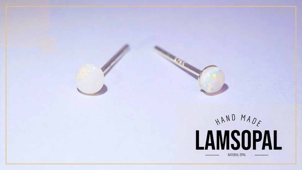 A pair of Lamsopal stud earrings featuring round white natural opal gemstones on silver posts, displayed on a light lavender background with a brand logo overlay in the bottom right corner.