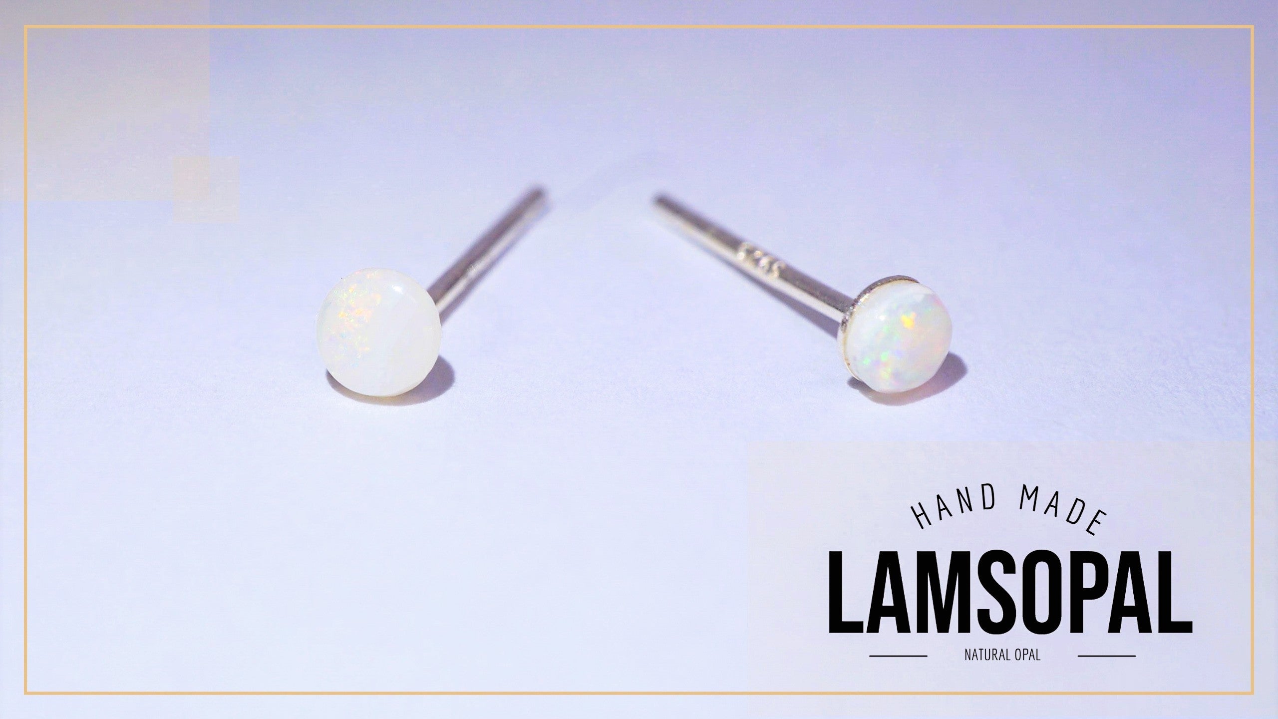 A pair of Lamsopal stud earrings featuring round white natural opal gemstones on silver posts, displayed on a light lavender background with a brand logo overlay in the bottom right corner.