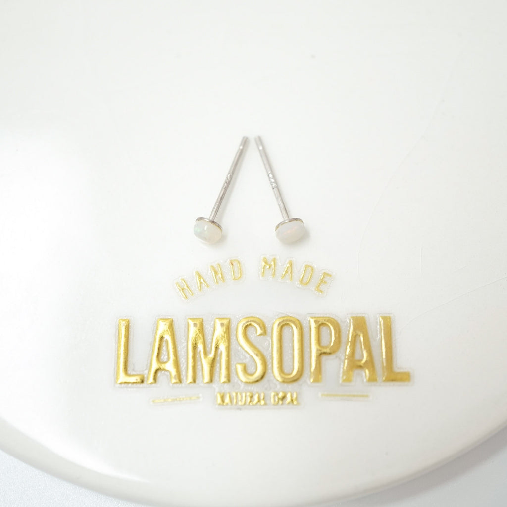 A pair of silver stud earrings with white oval inlays, displayed on a white circular surface above gold foil text that reads 'HAND MADE LAMSOPAL NATURAL Opal'.