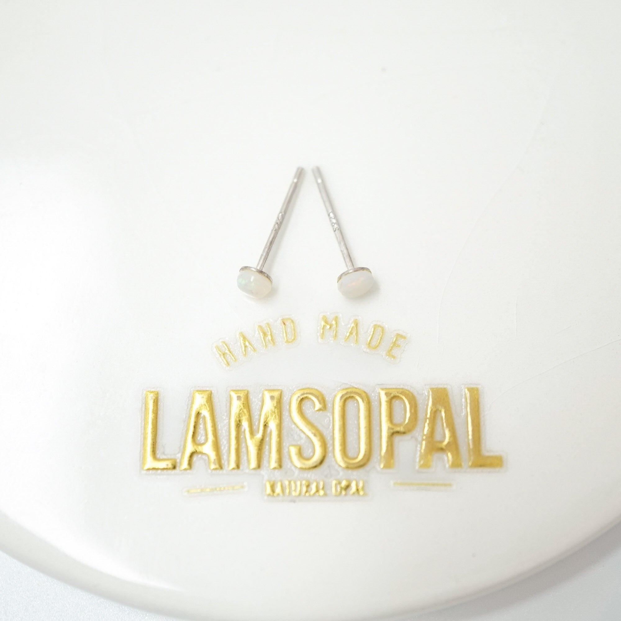 A pair of silver stud earrings with white oval inlays, displayed on a white circular surface above gold foil text that reads 'HAND MADE LAMSOPAL NATURAL Opal'.