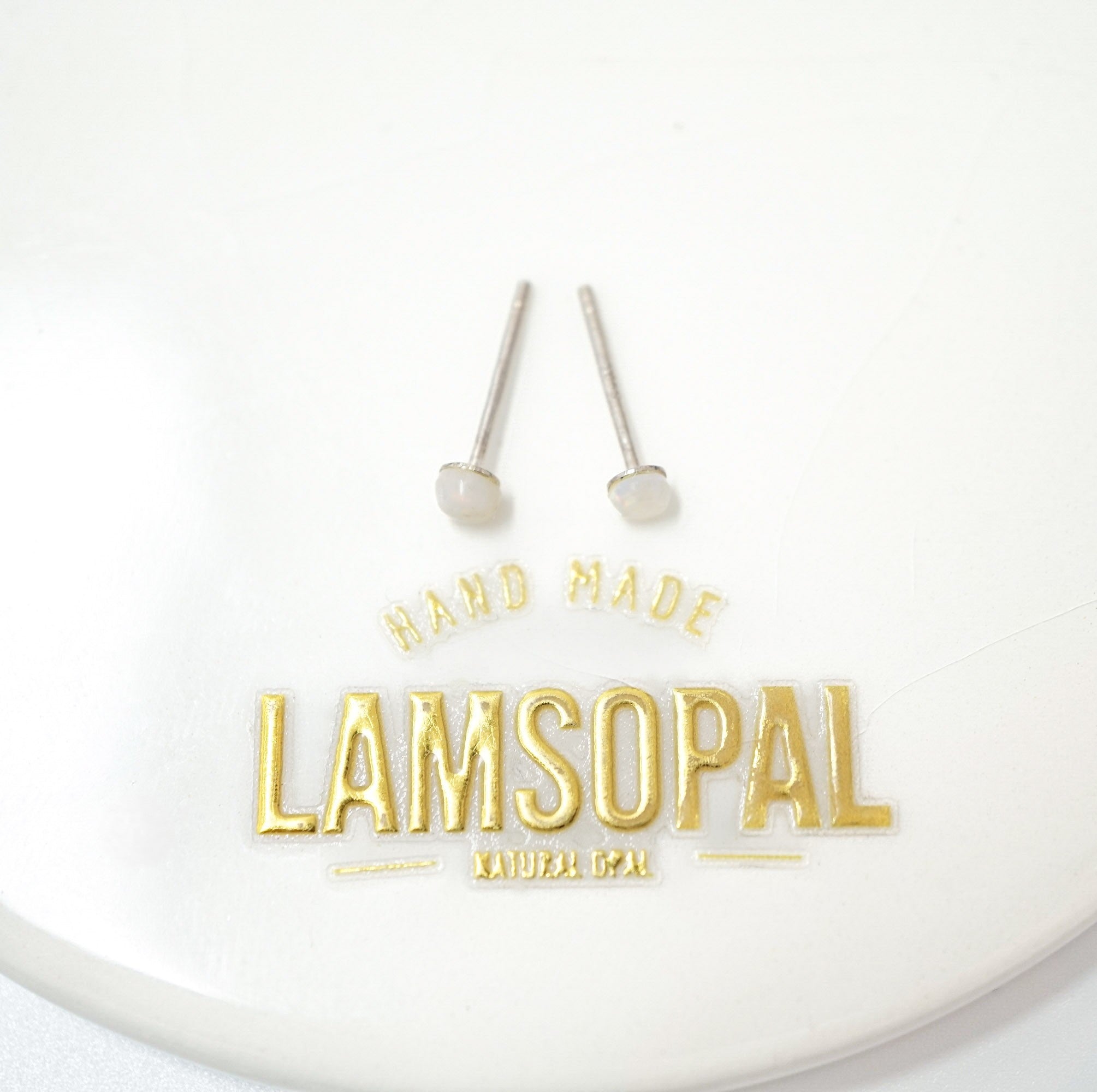 A pair of small silver stud earrings with oval clear stones, displayed above gold foil branding that reads 'HAND MADE LAMSOPAL NATURAL GMP' on a white background.