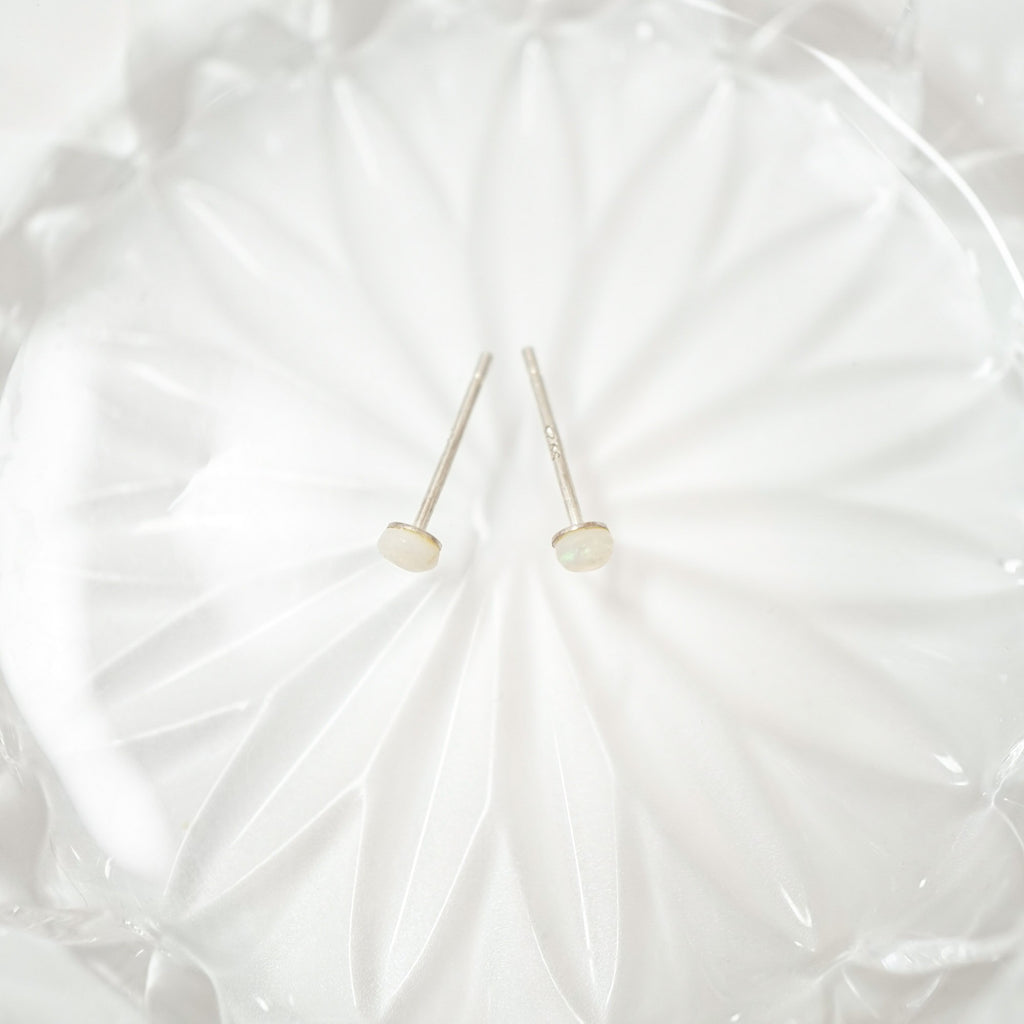 A pair of minimalist stud earrings with round, pale cream-colored cabochon stones on thin silver posts, displayed on a textured white glass dish.