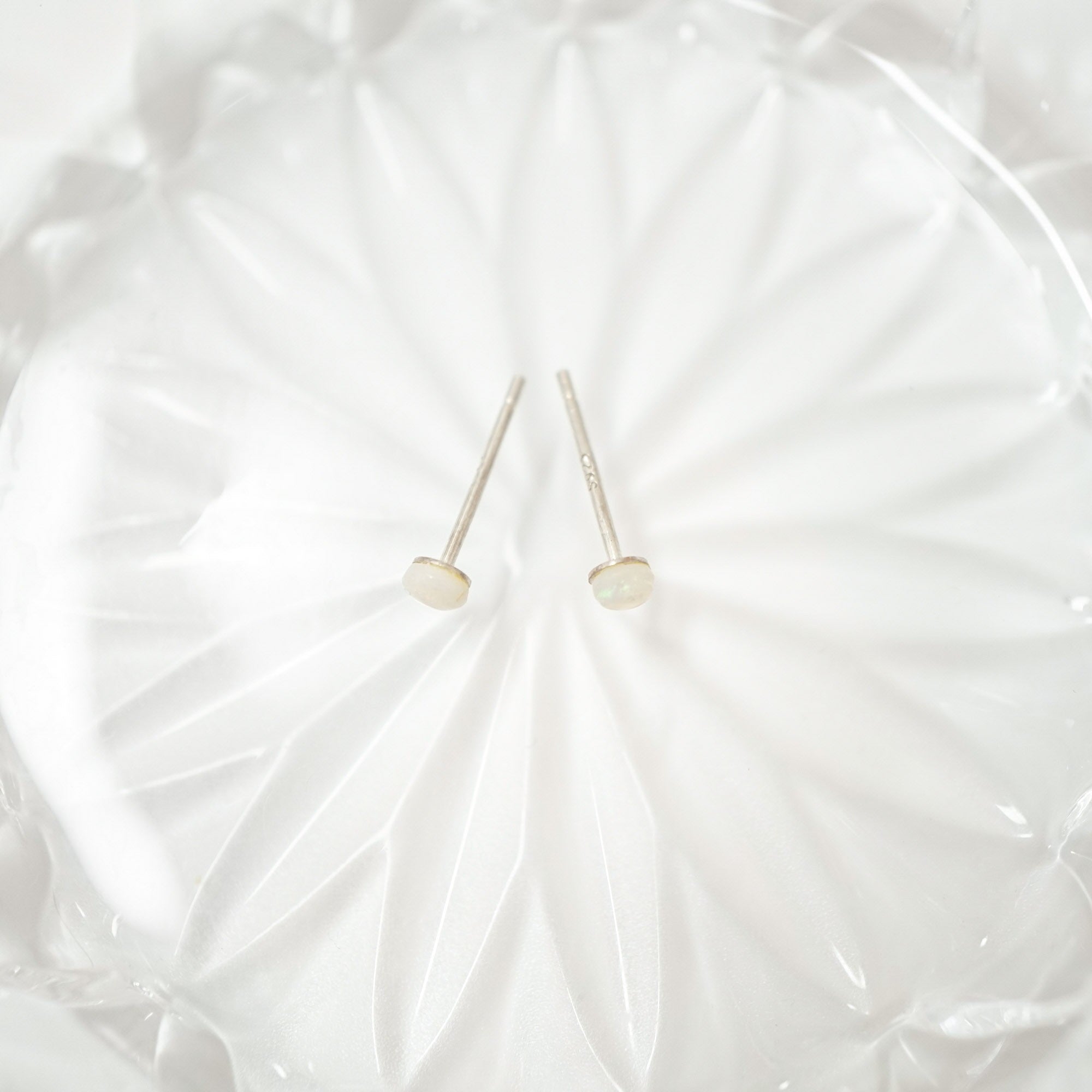 A pair of minimalist stud earrings with round, pale cream-colored cabochon stones on thin silver posts, displayed on a textured white glass dish.