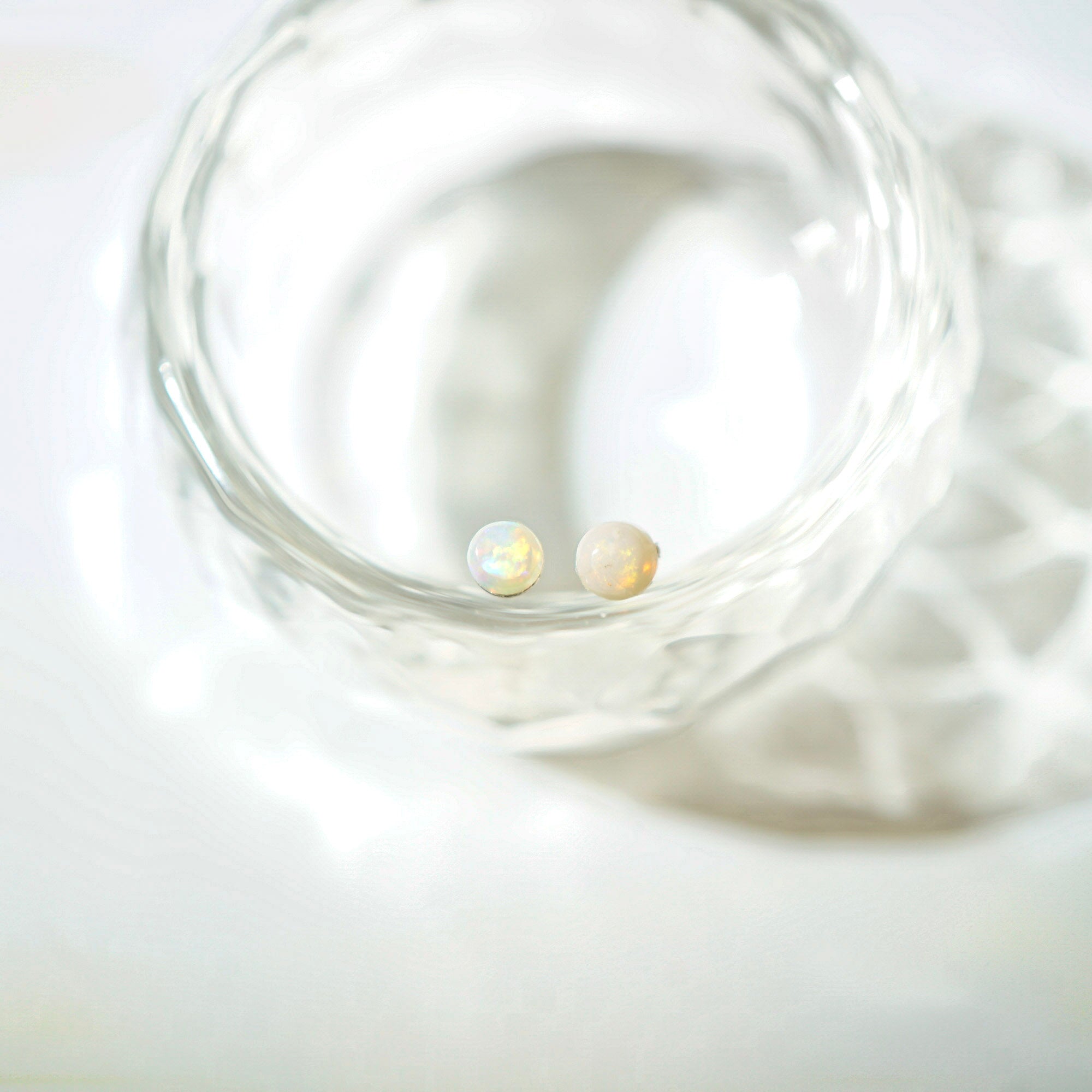 Two small iridescent pearl stud earrings resting inside a clear textured glass bowl on a soft white surface.