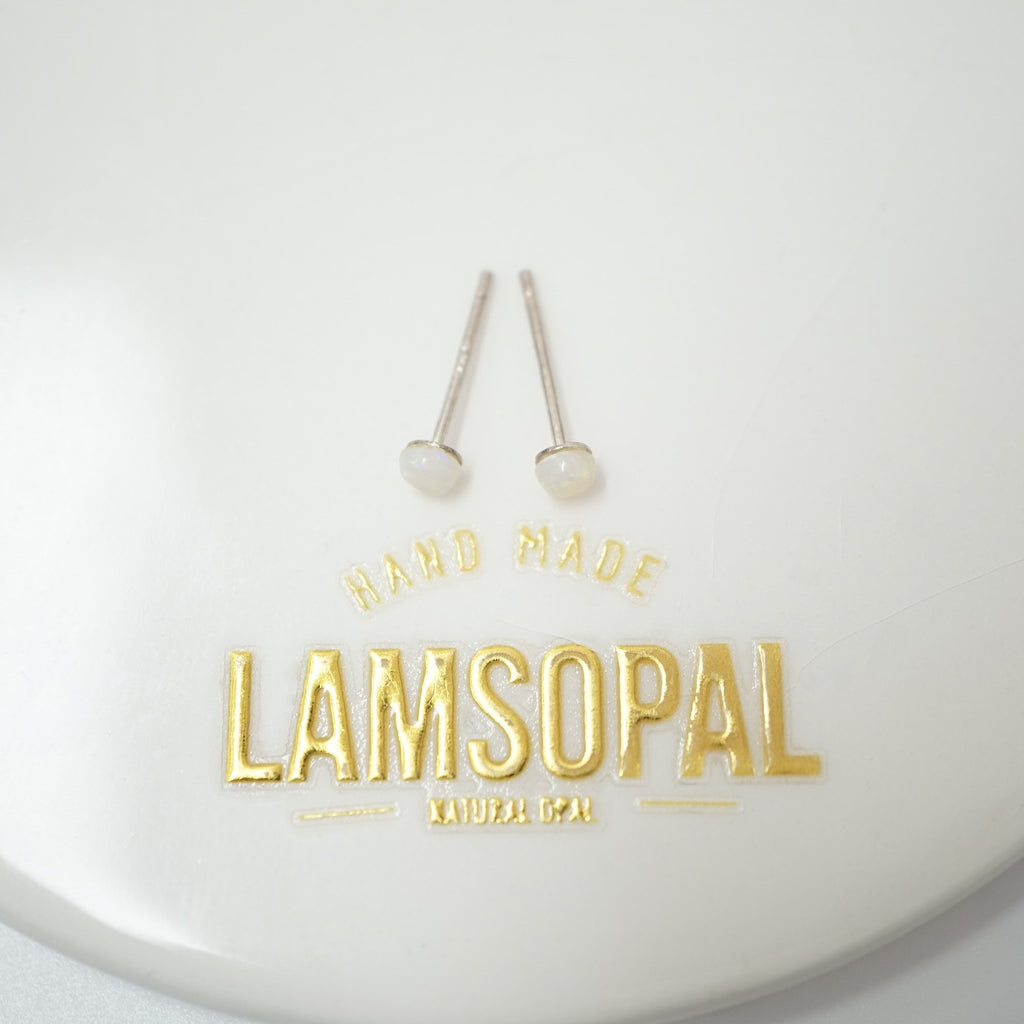 A pair of gold post earrings with round white bead-like studs, displayed on a white circular surface above gold foil branding that reads 'HAND MADE LAMSOPAL NATURAL GULAP'.