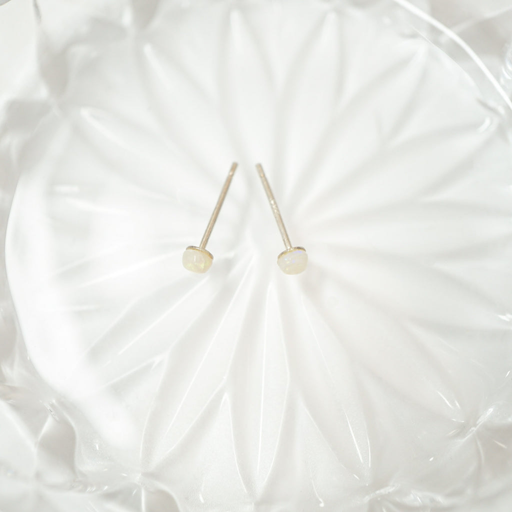 A pair of minimalist stud earrings with small, round, milky white beads on thin silver posts, displayed on a textured white glass plate.