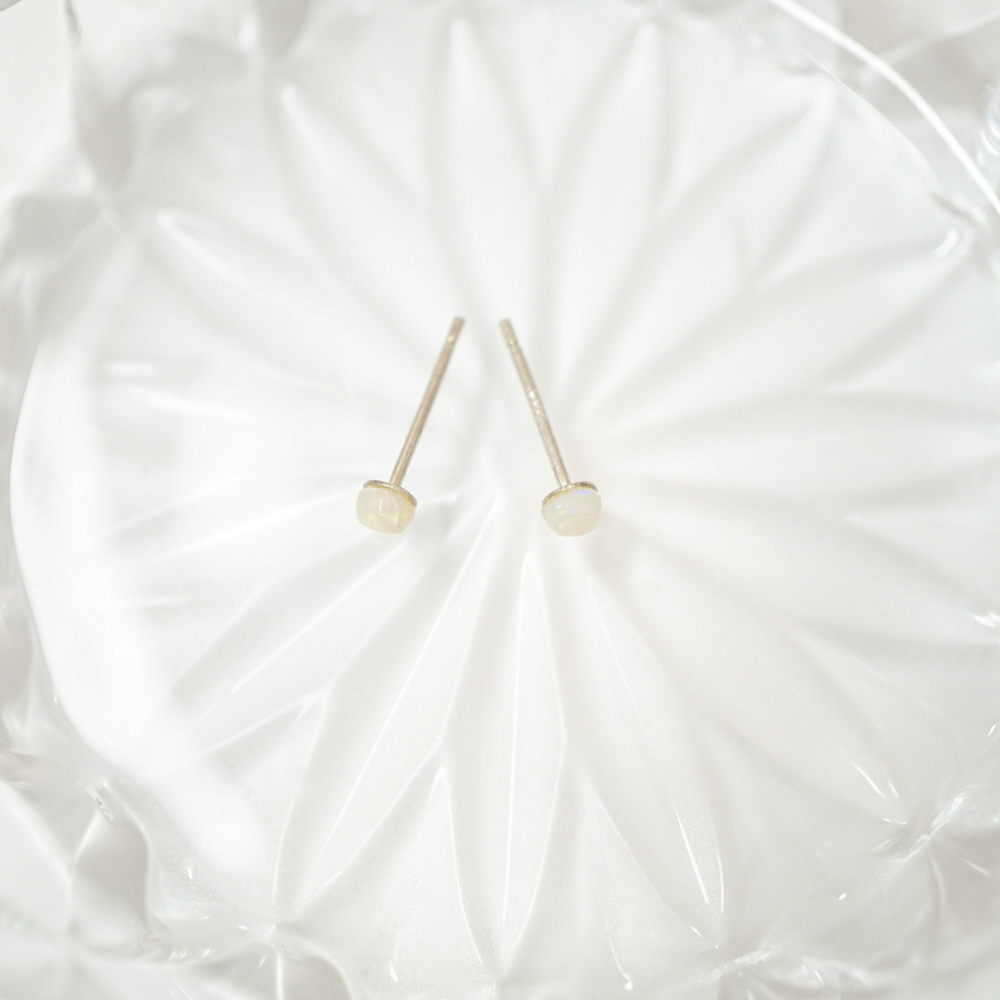 A pair of minimalist stud earrings with small, round, milky white beads on thin silver posts, displayed on a textured white glass plate.