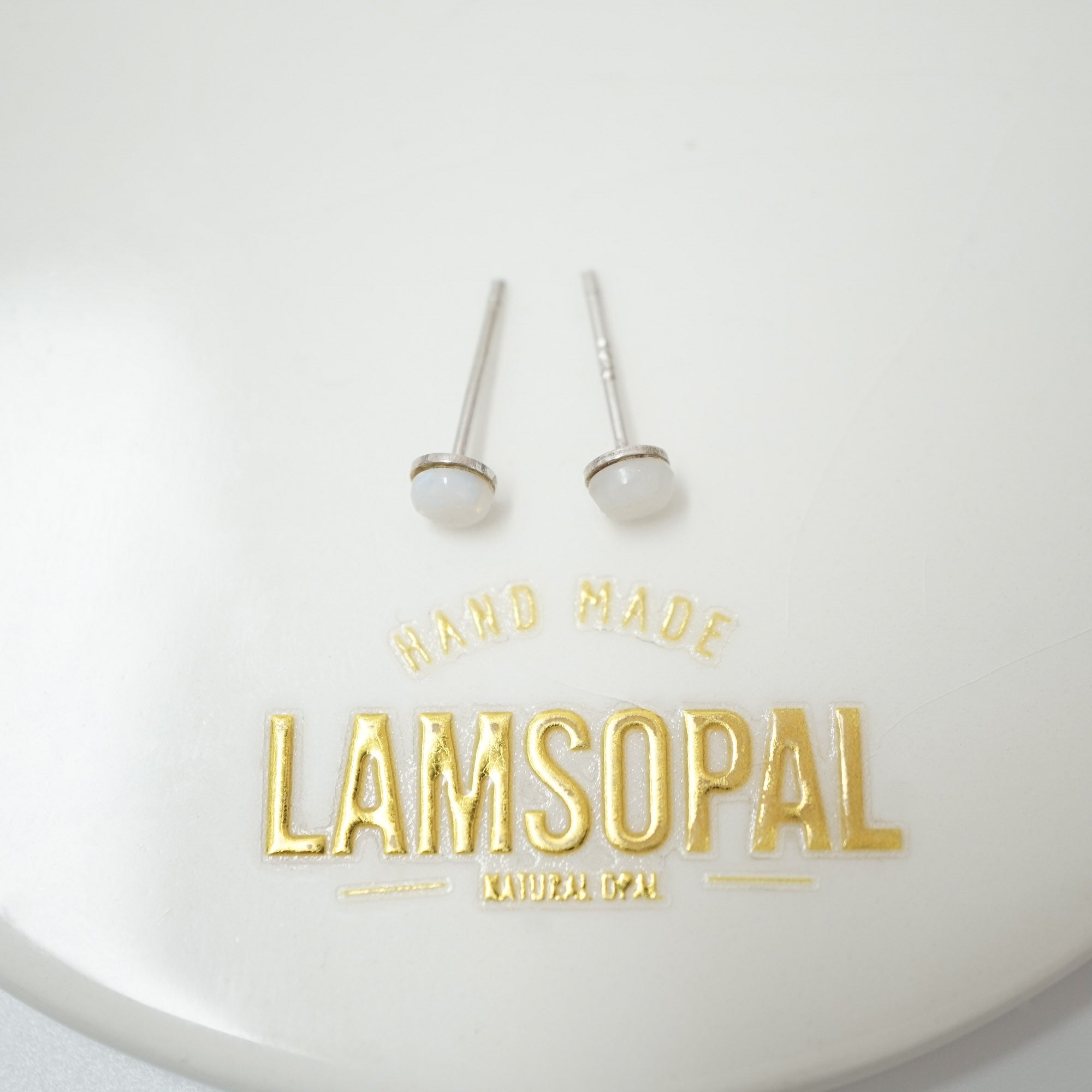 A pair of small silver stud earrings with oval white stone settings, displayed on a white surface above gold foil branding that reads 'HAND MADE LAMSOPAL NATURAL GEM'.