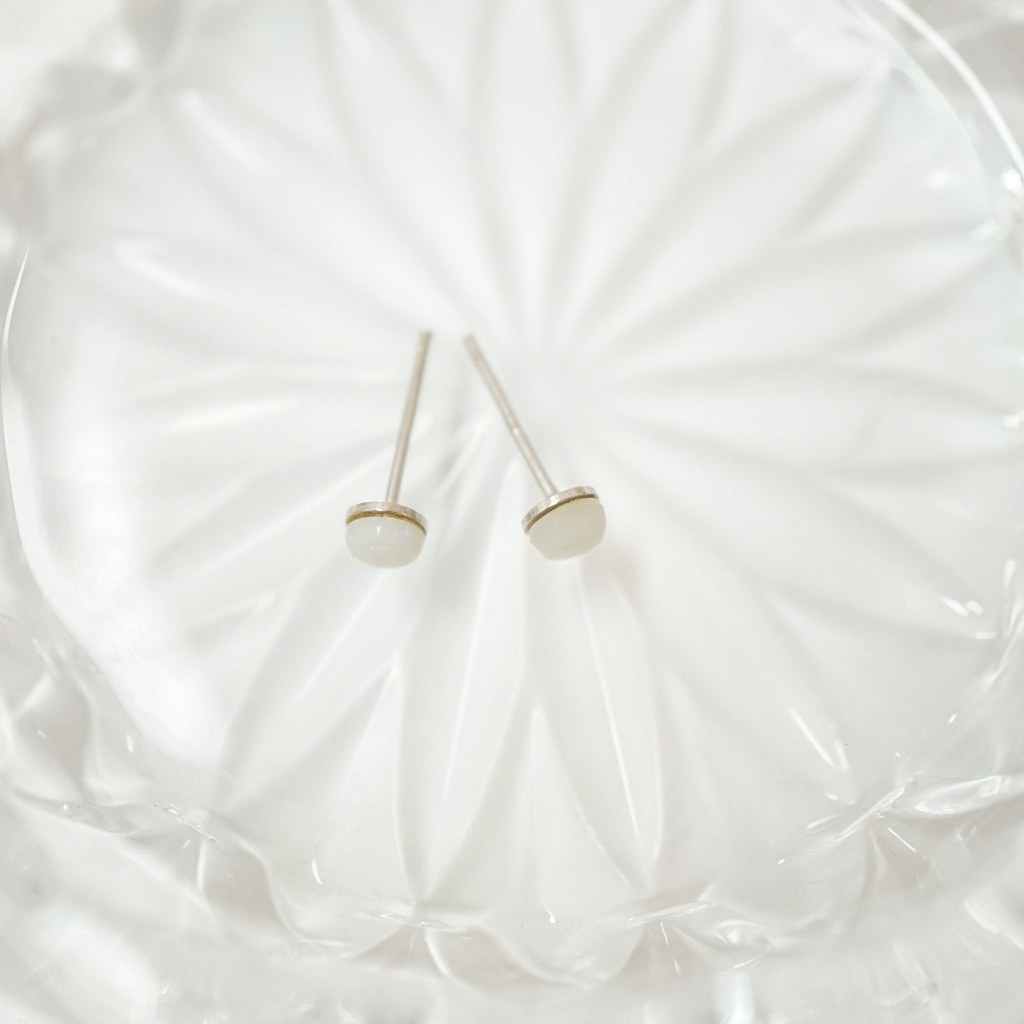 A pair of minimalist stud earrings with oval white inlays and thin gold-toned settings on silver posts, displayed on a textured white glass plate.