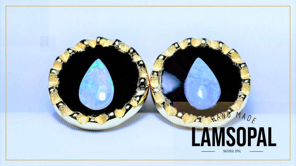 A pair of handmade Lamsopal natural opal teardrop stud earrings with gold bezel settings and black inlay, displayed on a light blue background with a gold border.