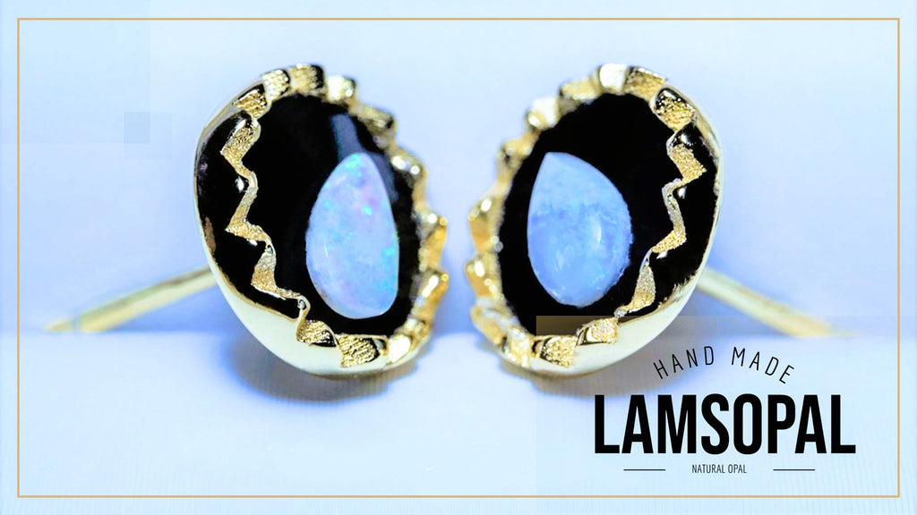 A pair of handmade Lamsopal stud earrings featuring natural blue opal centers set in gold bezel settings with black resin borders, shown against a light blue background with a brand logo overlay.