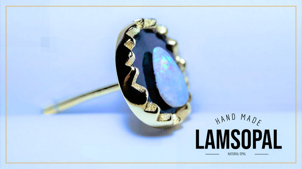 A gold ring featuring a round natural opal gemstone with iridescent blue, white, and black play-of-color, set in a scalloped gold bezel. The ring is displayed on a light blue background with a gold border, and the text 'HAND MADE LAMSOPAL' is visible in the lower right corner.