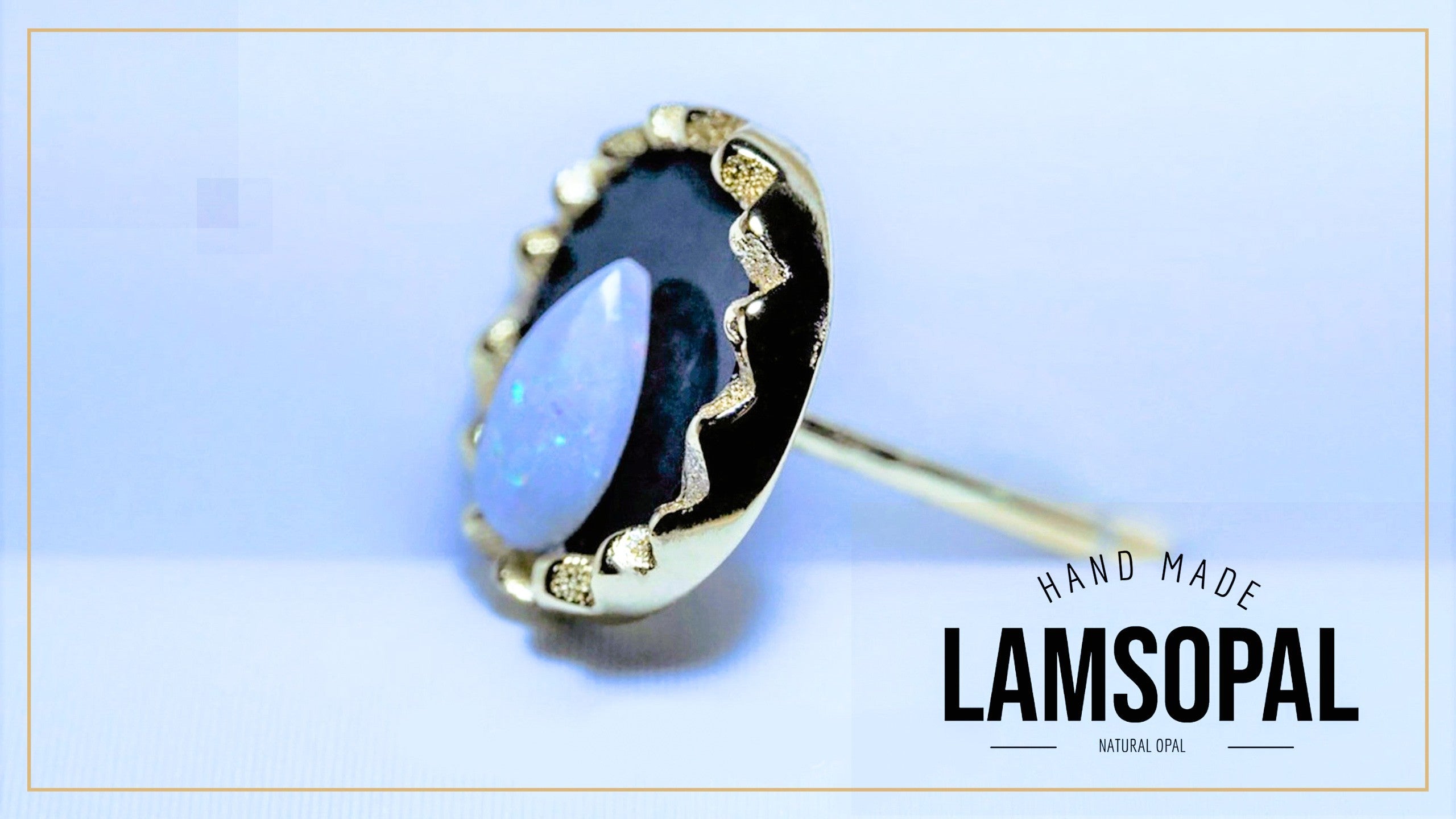 A single Lamsopal hand-made ring featuring a natural opal with a blue-white center and a dark blue outer edge, set in a gold-toned bezel with small gold inlays. The ring is shown on a light blue background with a gold border, and the brand name 'LAMSOPAL' is visible in the bottom right corner.