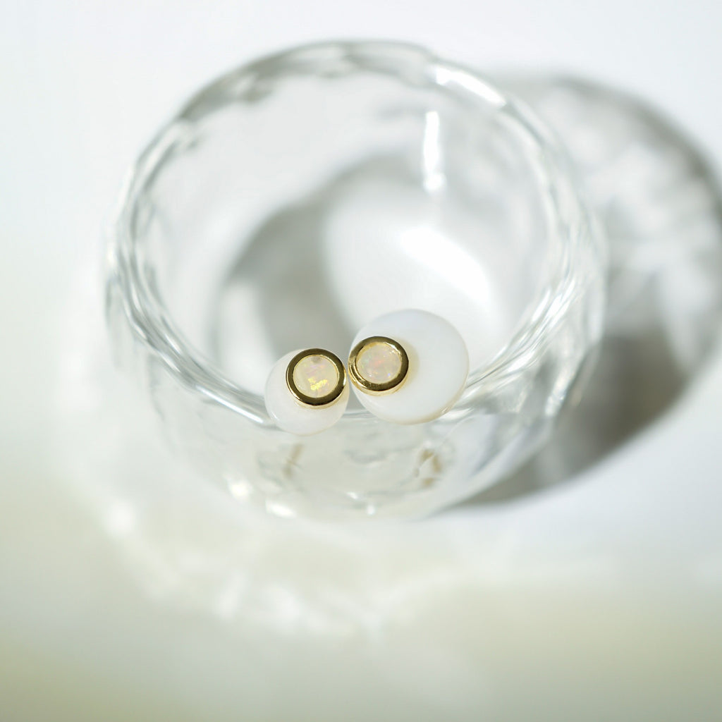 A pair of small stud earrings with round white stones and thin gold settings, resting inside a clear glass dish on a soft white surface.